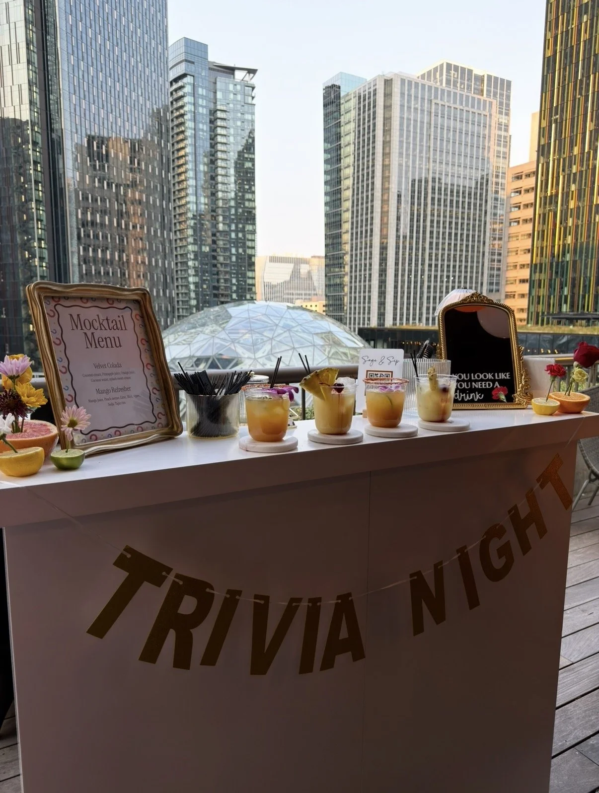 A white table with four cocktails topped with fruit, a framed mocktail menu and a small blackboard sign, decorated with flowers, set against a city skyline with tall buildings.