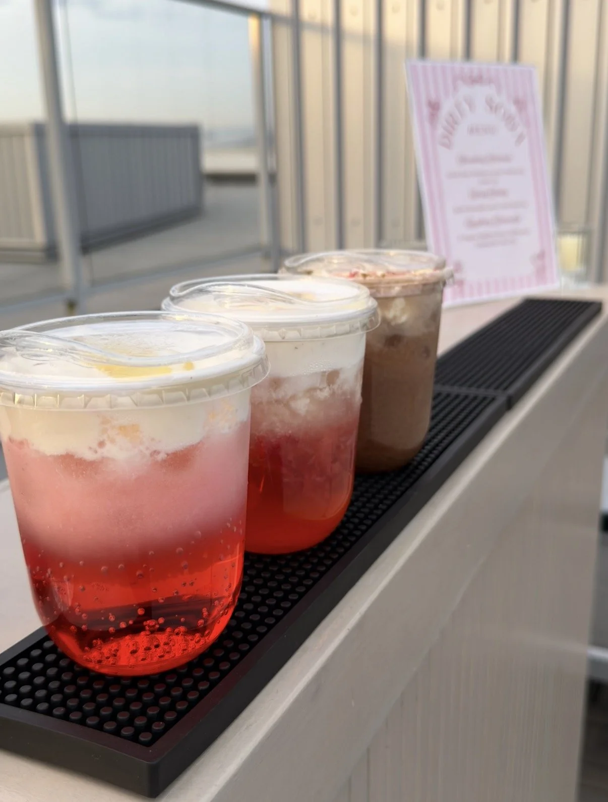 Three plastic cups with layered pink, white, and brown dirty soda drinks, topped with cold foam, lined up on a black rubber mat with a blurred pink and white sign in the background.