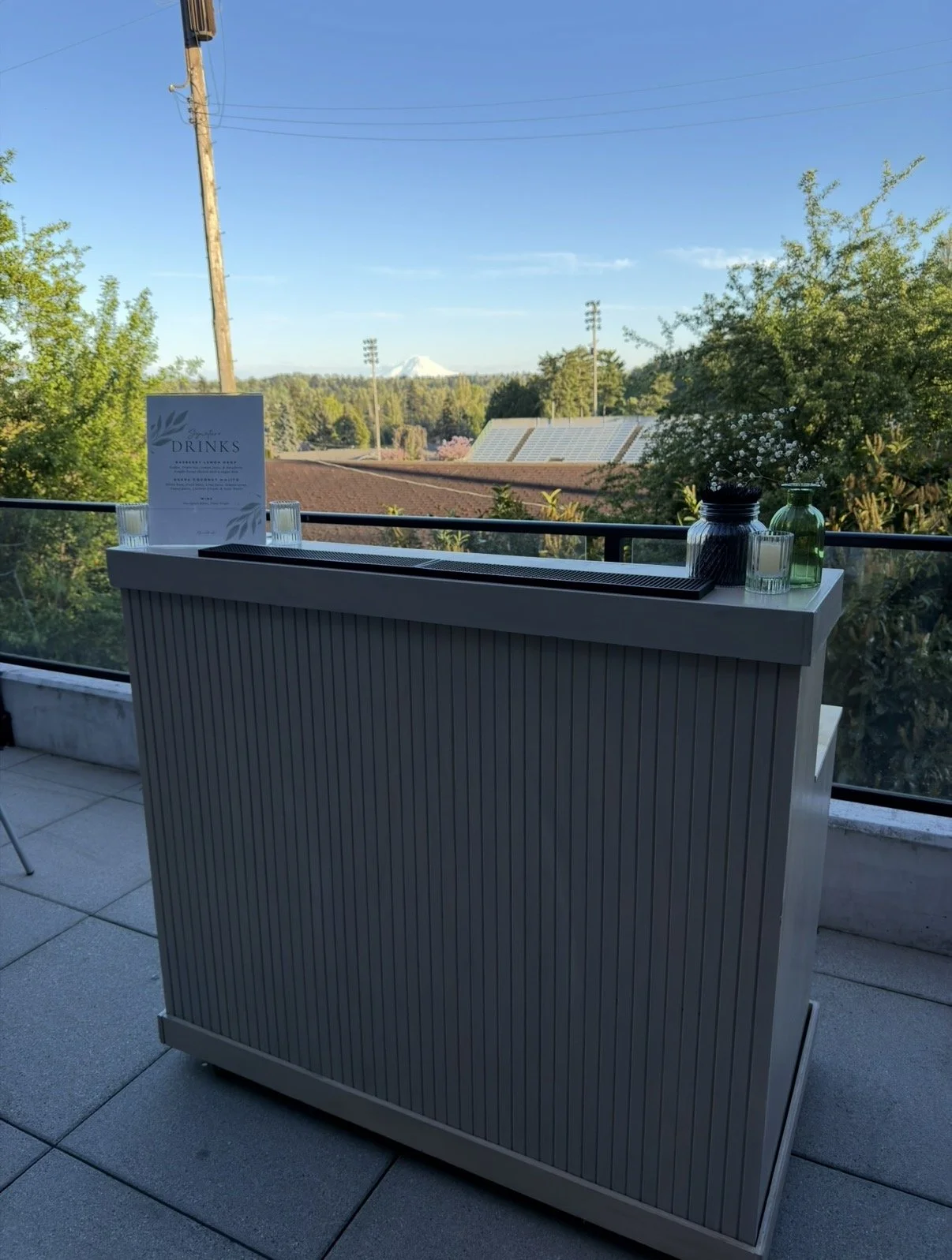 Outdoor balcony with a modern bar or counter, decorative vases, a menu, and scenic view of trees and distant mountain in the background.