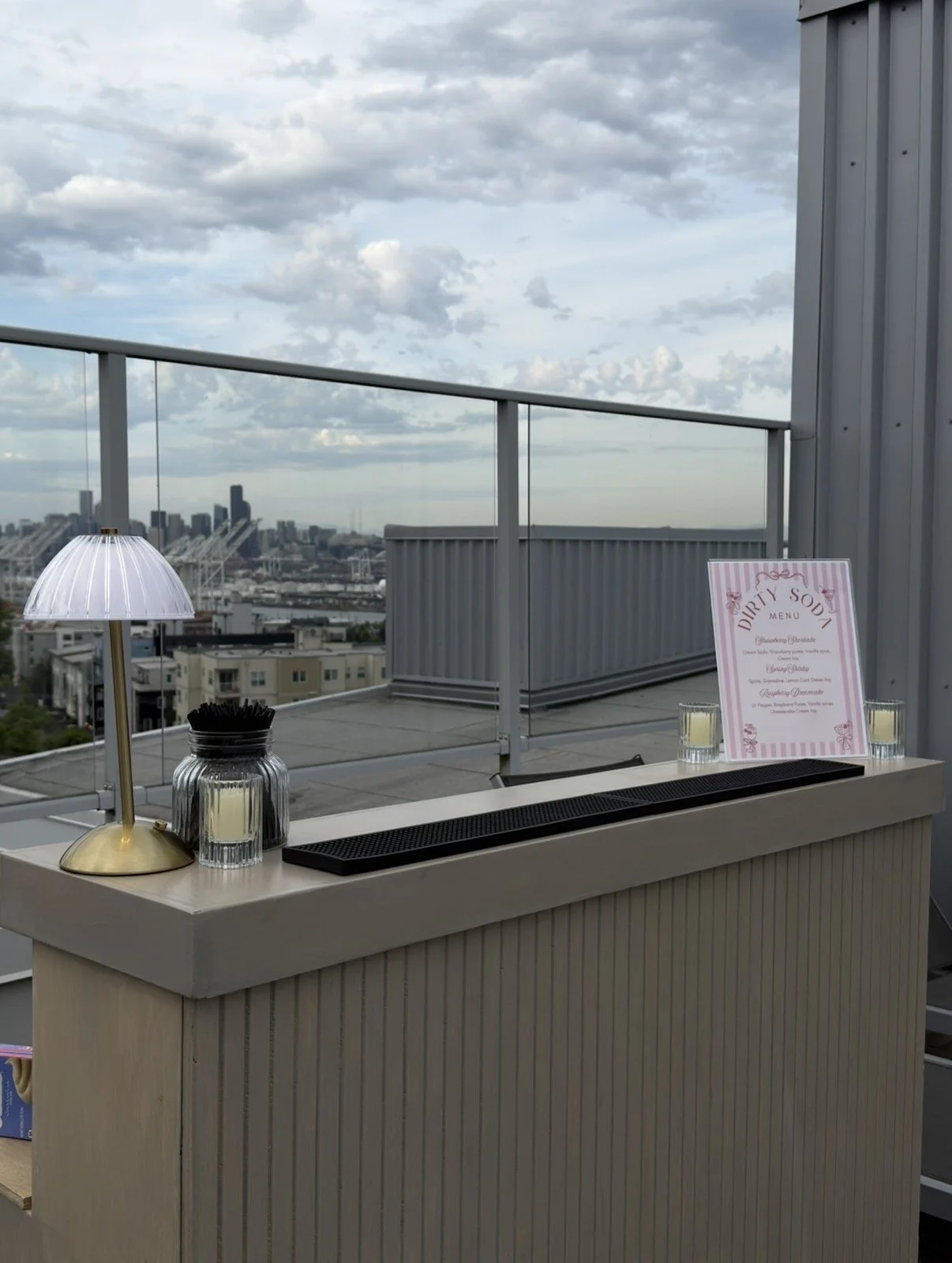 A rooftop bar setup with an aesthetically pleasing mobile bar, a white lamp with a gold base, two glasses with candles, a jar of black stirrers, and a menu with pink accents. The city skyline and cloudy sky are visible in the background.