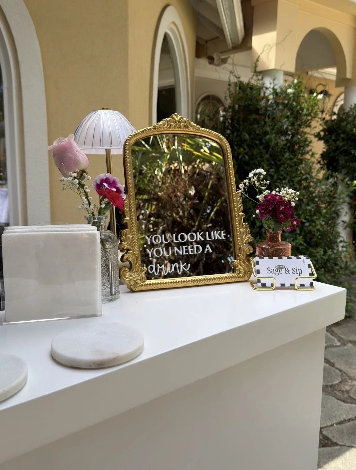 A decorated table with a gold-framed mirror that has a sign saying "You look like you need a drink." There are small flower arrangements in glass bottles, a lamp, and a sign labeled "Sage & Sip."