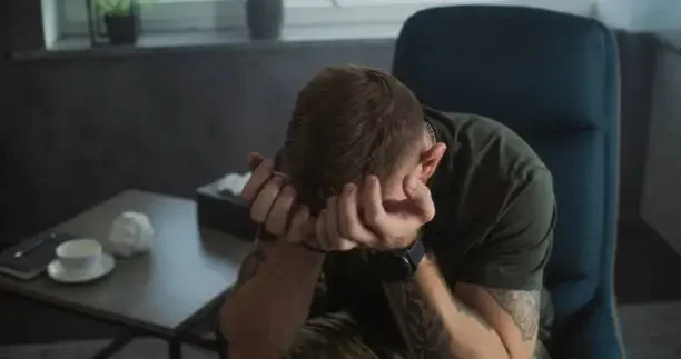 A young man sitting in a chair with his head resting on his hands, looking down, in an office or room setting.