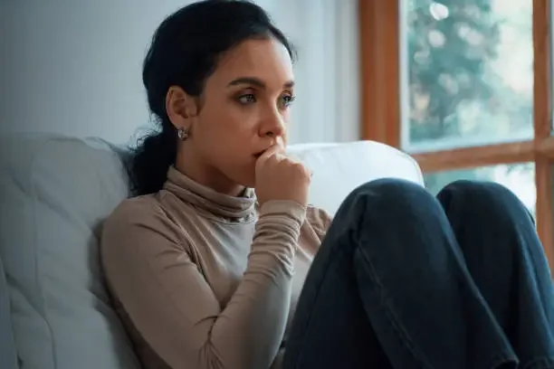 A woman sitting on a couch near a window, wearing a beige turtleneck sweater and blue jeans, with her hand near her mouth as if thinking or worried.
