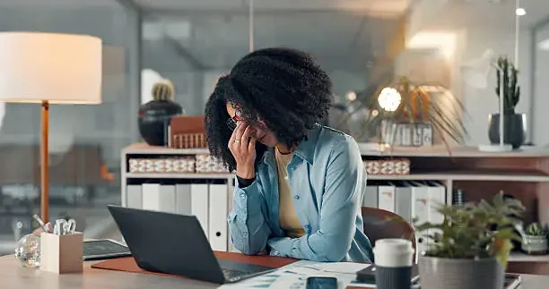 A woman sitting at a desk in an office, holding her forehead and appearing stressed, with a laptop and coffee cup on the desk.