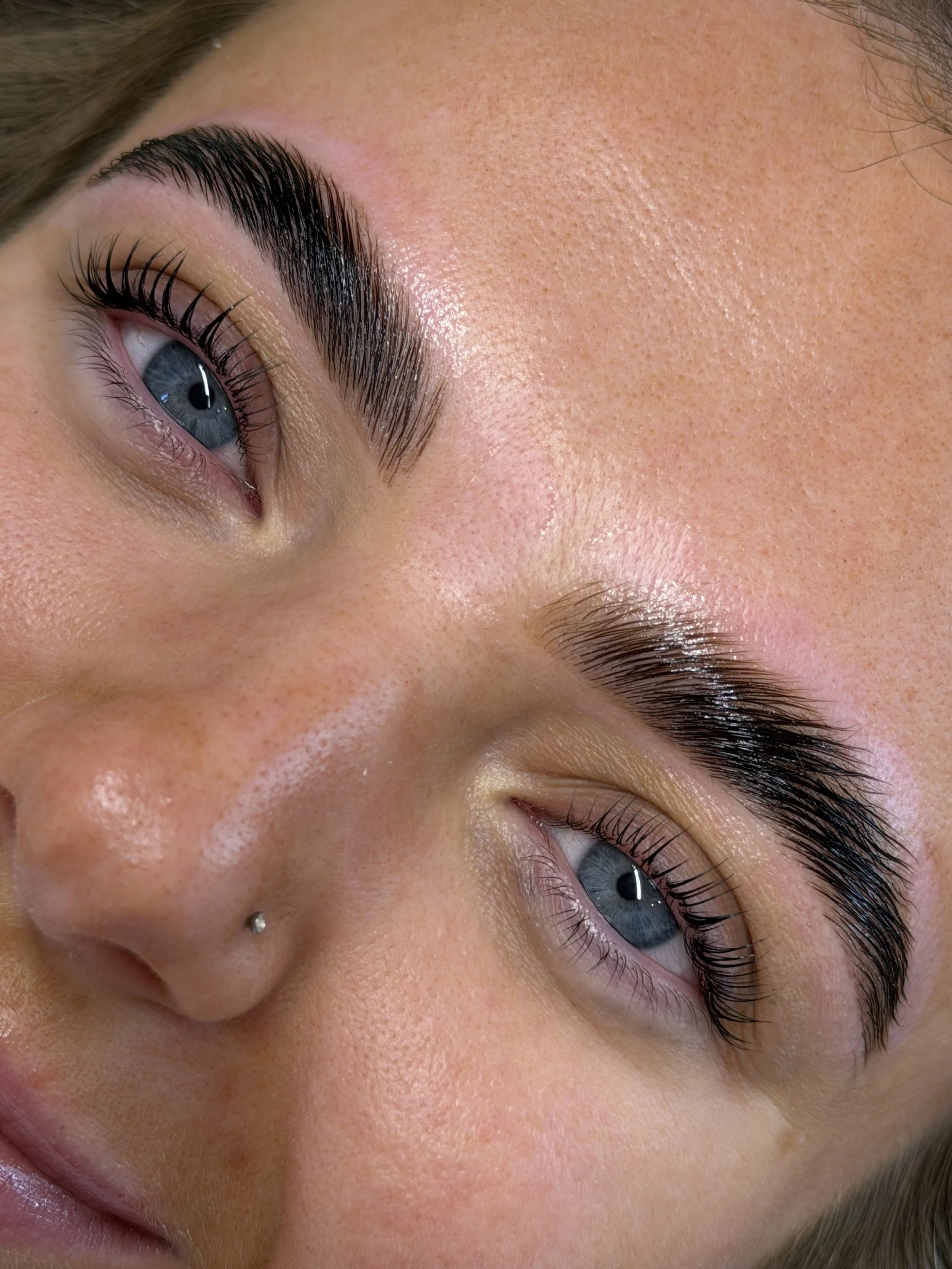 Close-up of a person's eye, with thick, well-groomed eyebrows, long eyelashes, and blue irises, along with a small nose piercing.