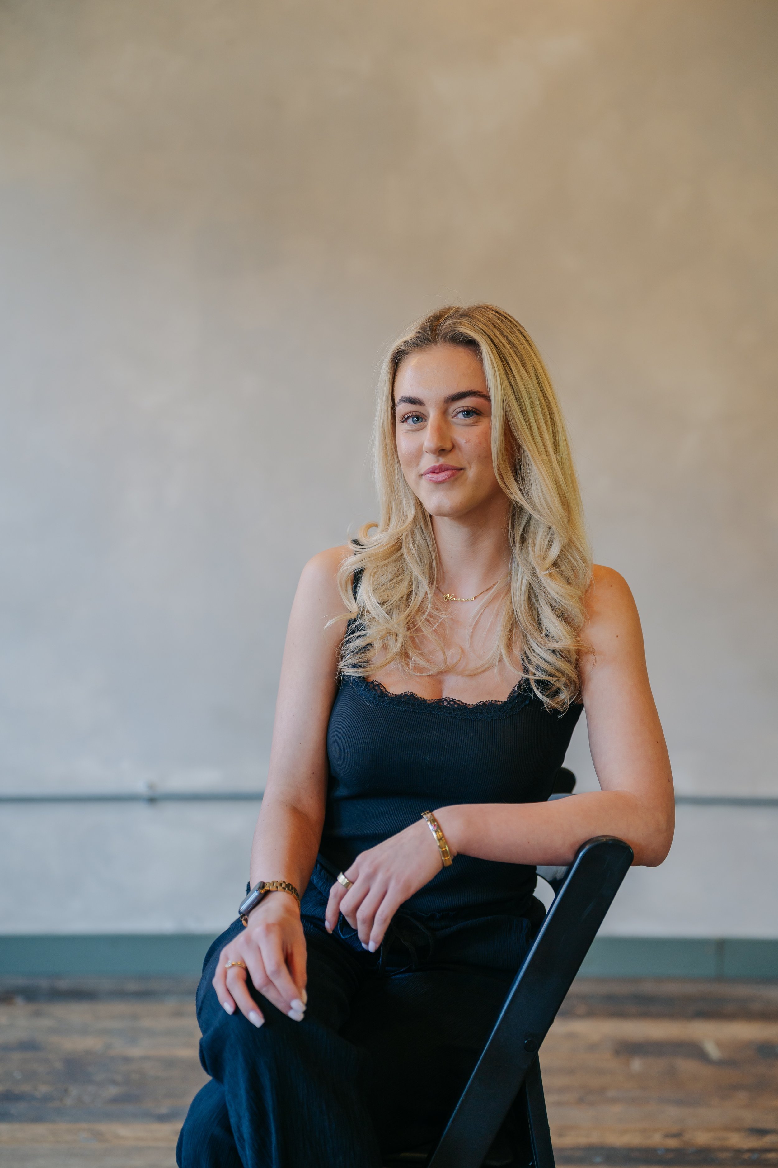 A young woman with blonde hair sitting on a black chair against a plain, light-colored wall. She is wearing a black top and gold jewelry, looking at the camera with a slight smile.