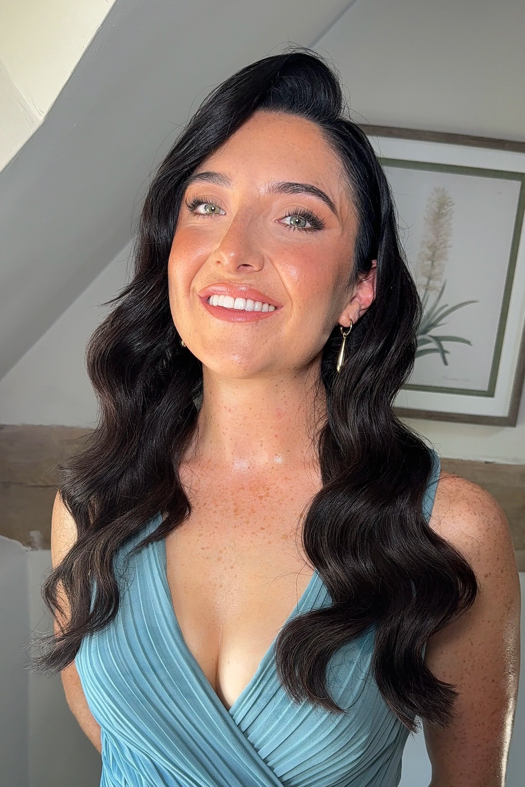 Close-up of a woman with long, wavy black hair, light green eyes, wearing a light blue sleeveless top with pleats, earrings, and makeup with mascara and pink lipstick, standing in front of a framed botanical print.