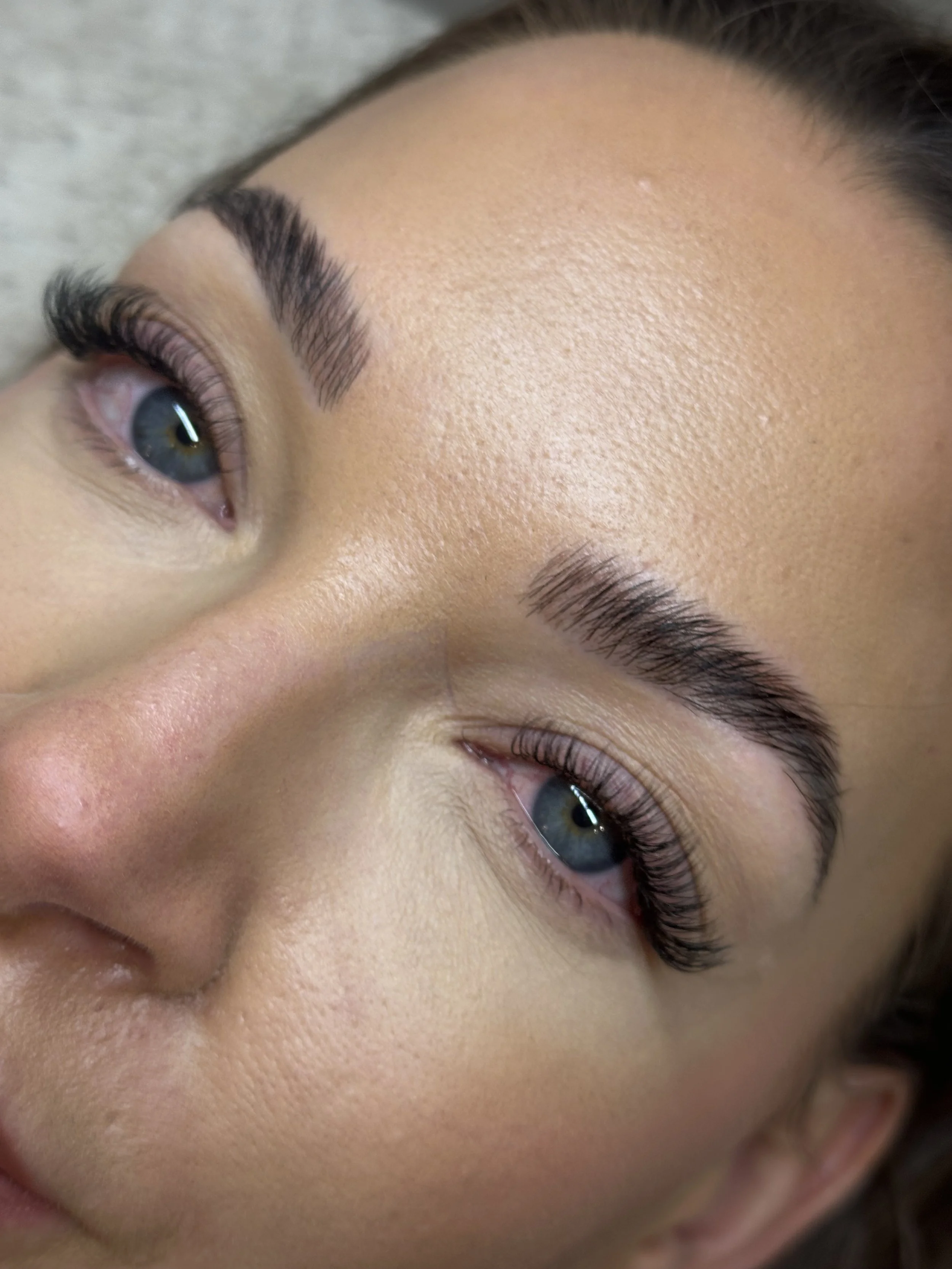 Close-up of a woman's face focusing on her eye, eyebrow, and forehead, showcasing well-groomed eyebrows, long eyelashes, and smooth, glowing skin.