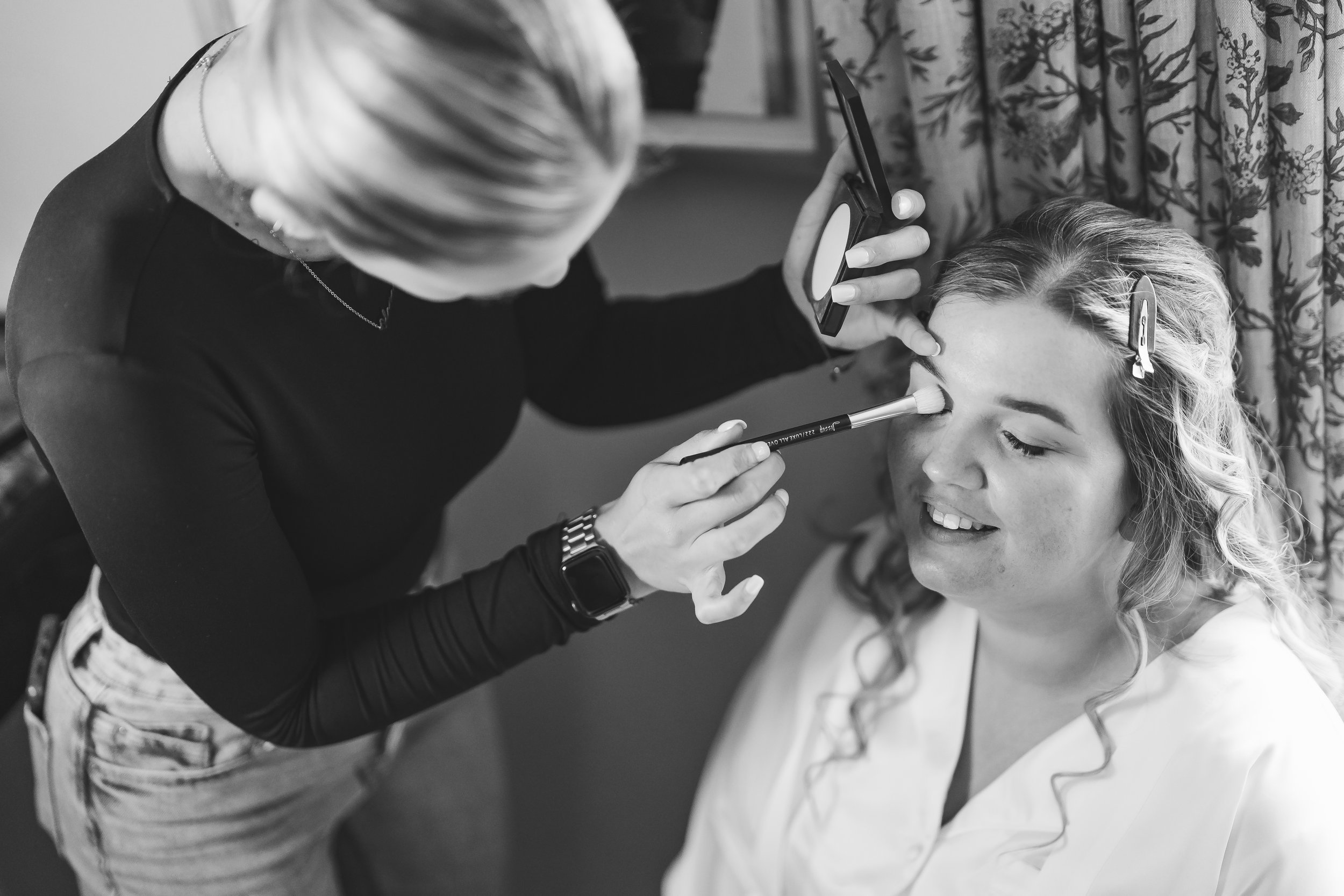 A woman with blonde hair getting her makeup done by a makeup artist in a room with floral curtains