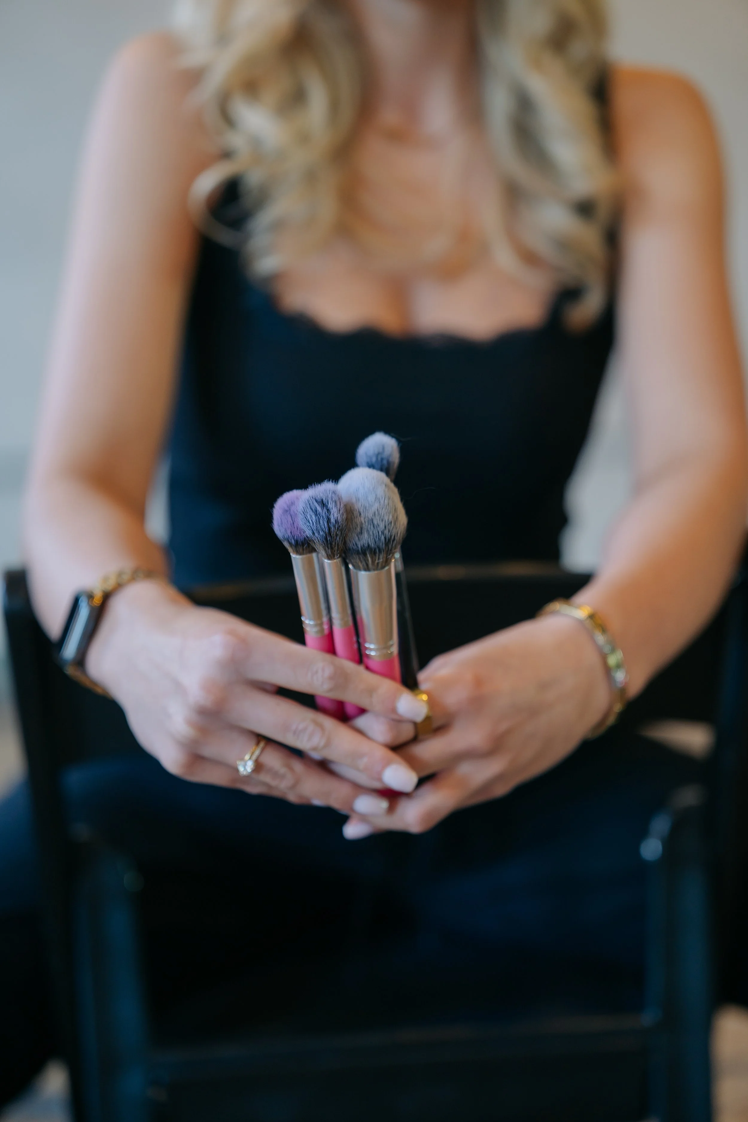 A woman holding makeup brushes in her hands, sitting on a chair, wearing a black sleeveless top, with blonde curly hair, in an indoor setting.