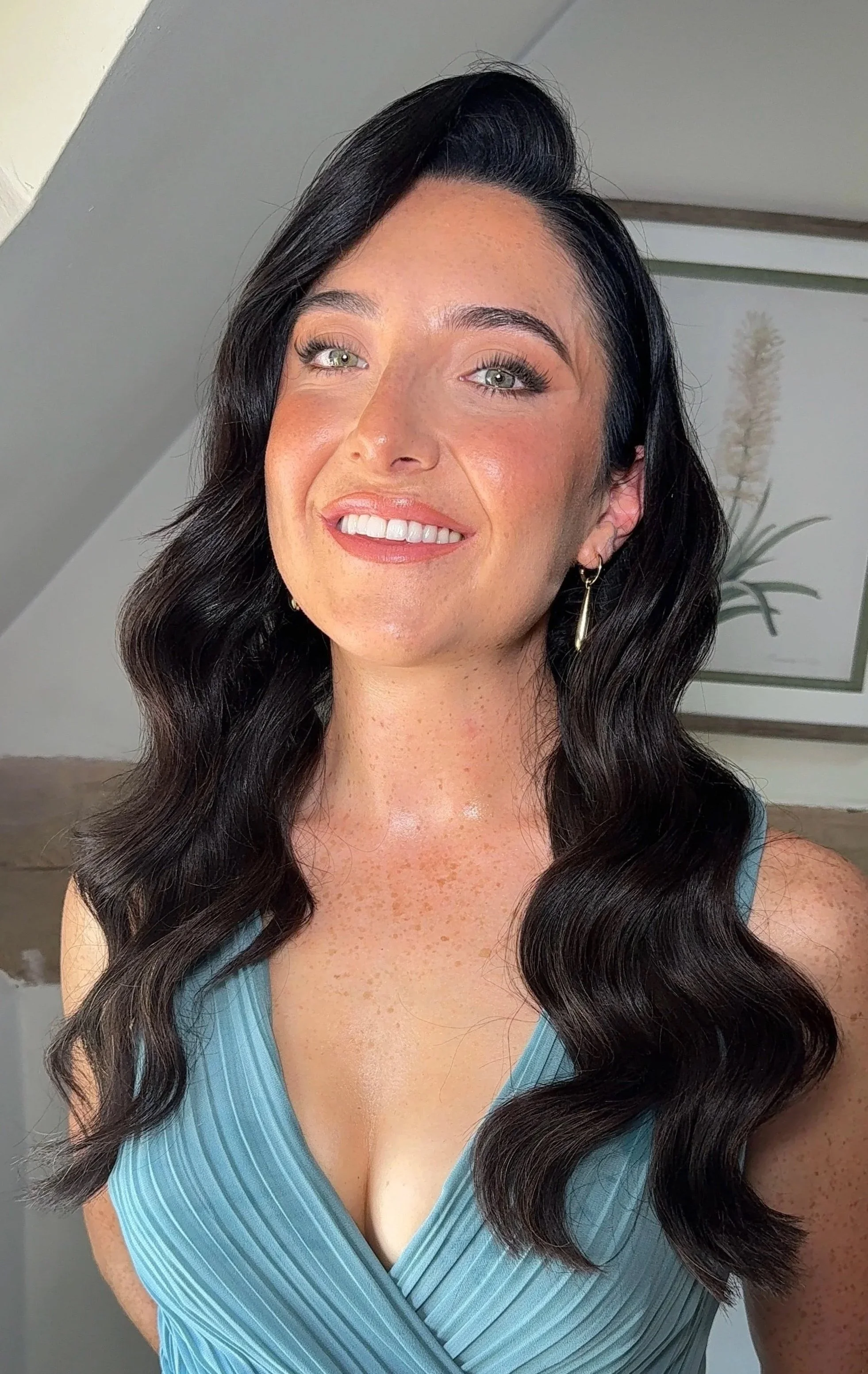 Close-up of a woman with long, wavy black hair, wearing a blue sleeveless dress with a V-neck, smiling with white teeth, wearing gold earrings, with a background featuring a potted plant and a wall.