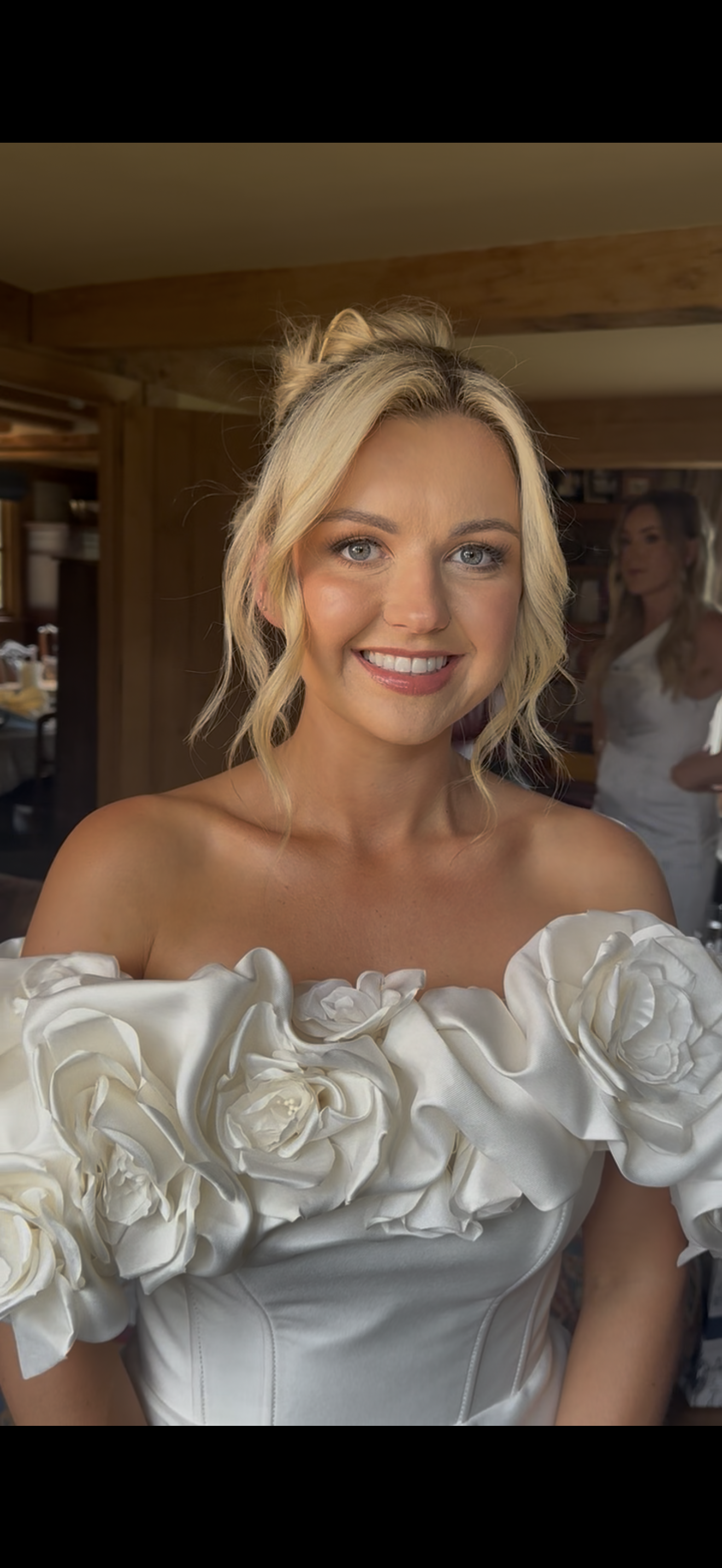 A smiling woman with blonde hair in an off-the-shoulder white dress with floral ruffles, indoors with wooden walls, another woman in white is in the background.