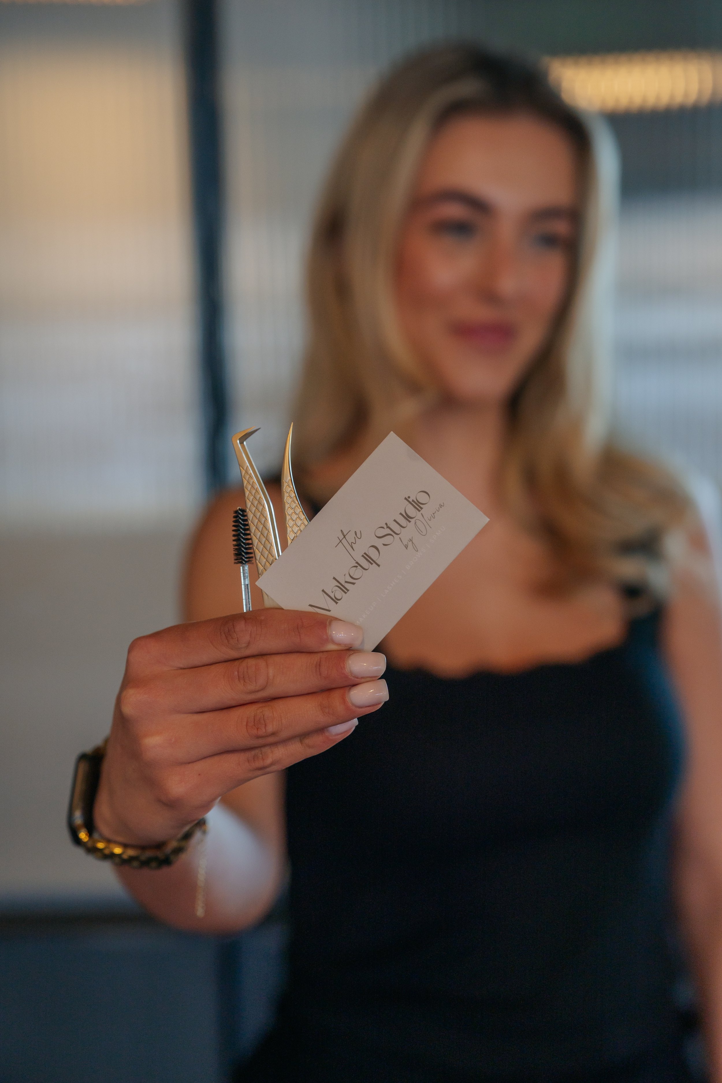 A woman with blonde hair holding a business card and beauty tools in an indoor setting.