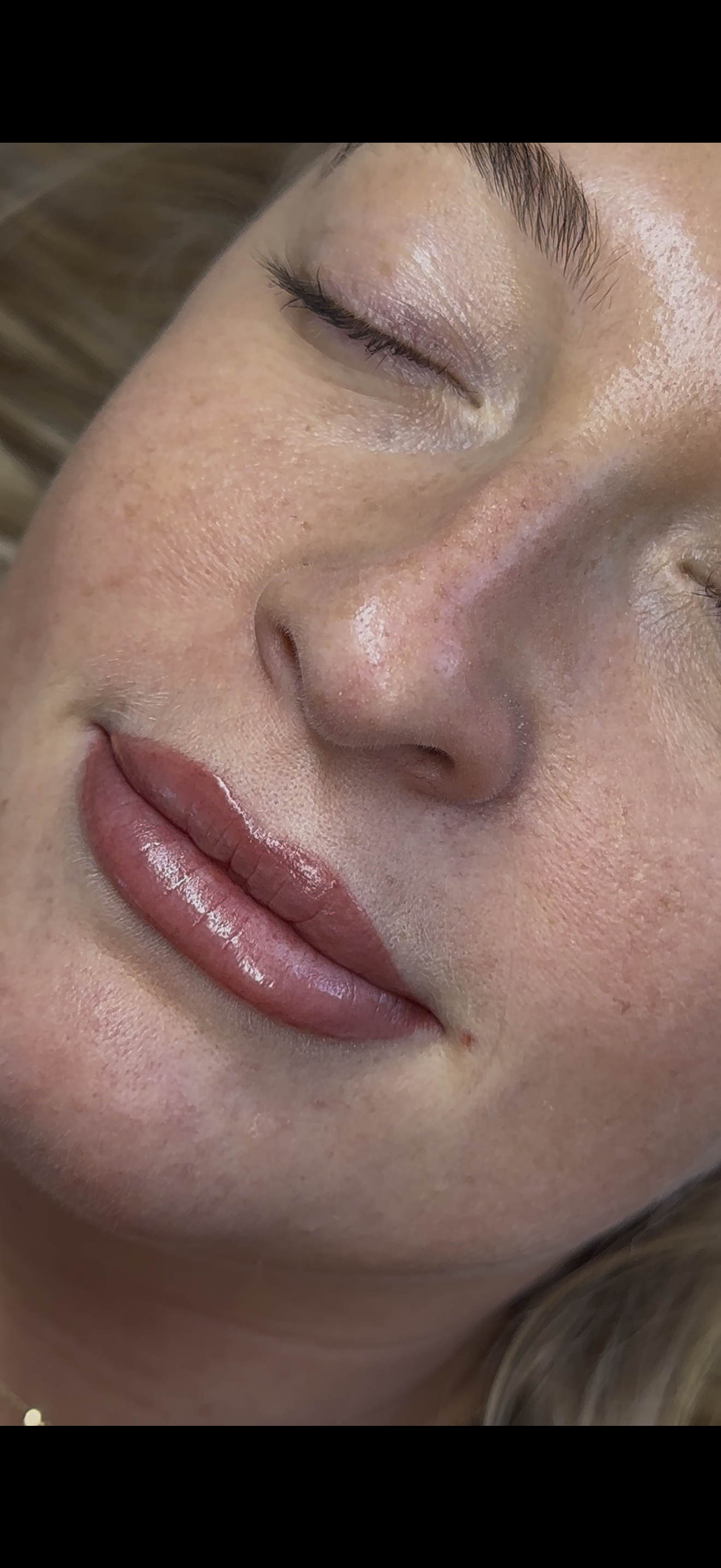 Close-up of a woman's face with her eyes closed, showing her lips with glossy lipstick, smooth skin, and well-groomed eyebrows.