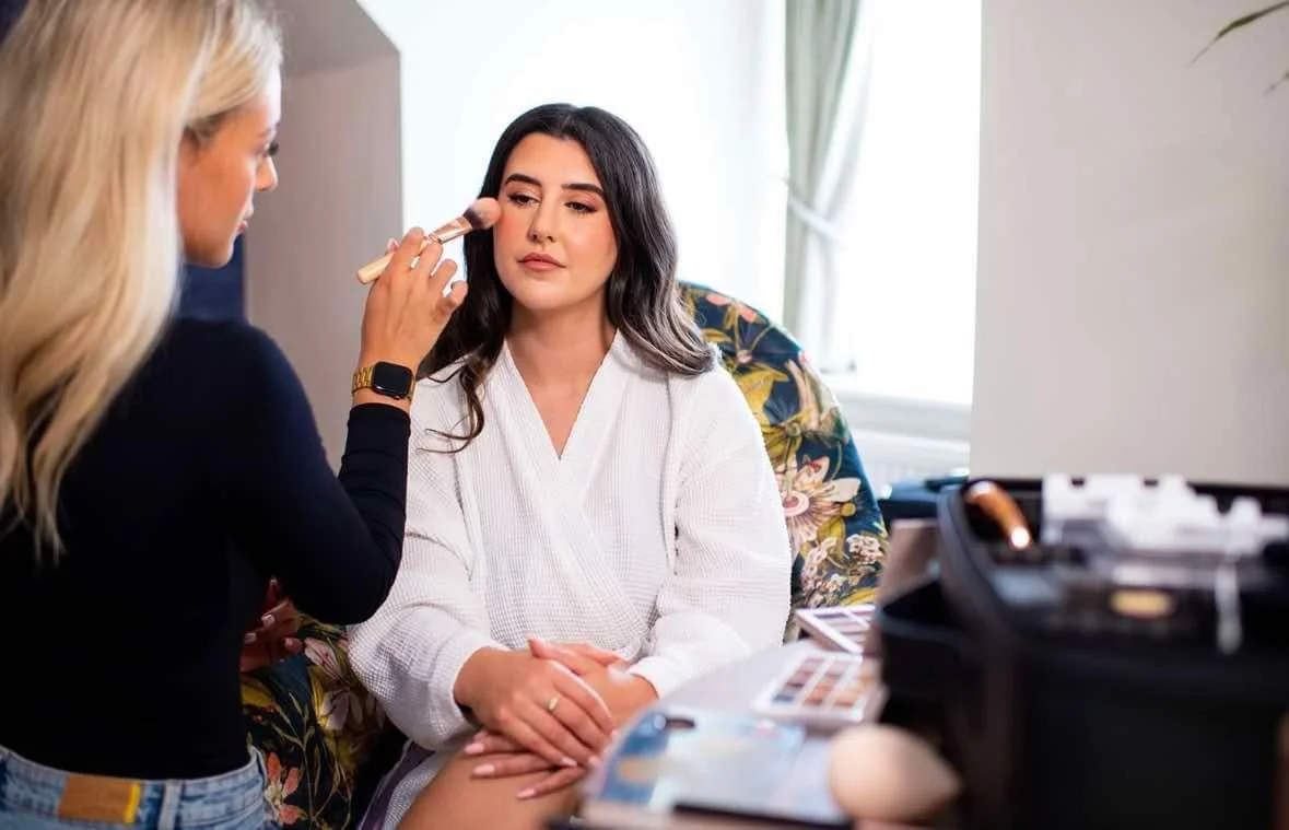A woman with dark hair sitting on a floral chair getting her makeup applied by a makeup artist with blonde hair.