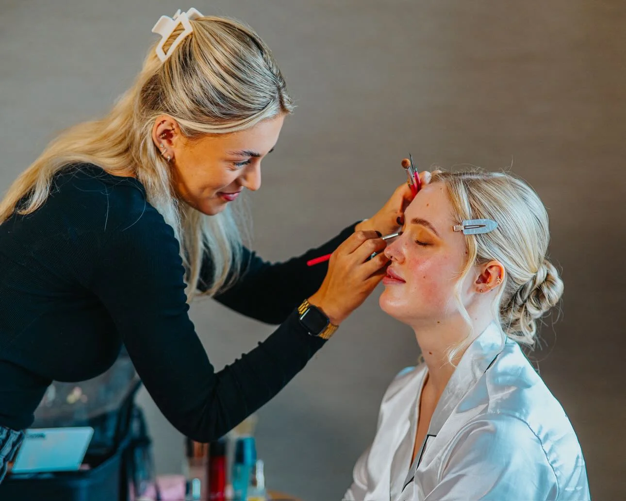 Makeup artist applying makeup to a woman with blonde hair seated with her eyes closed.