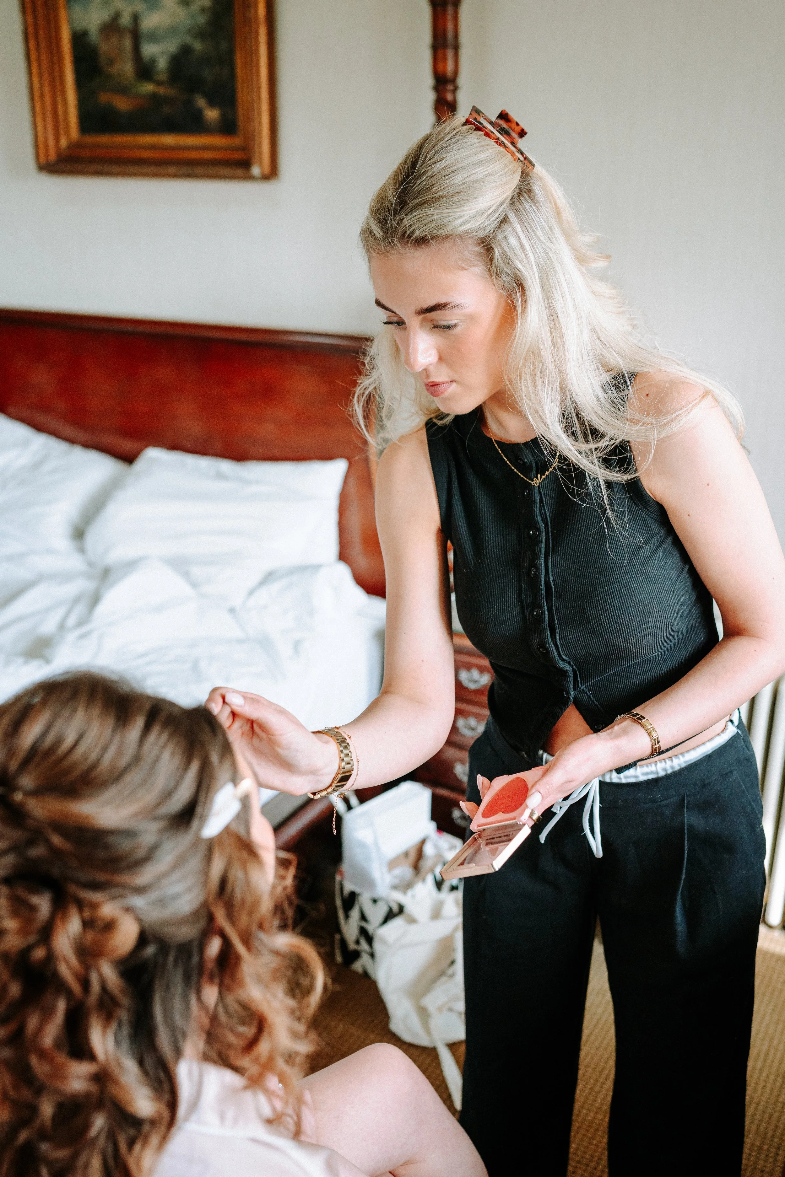 A young woman with platinum blonde hair and a tortoiseshell hair clip is applying makeup or skincare to another woman with curly, reddish-brown hair in a bedroom. The bed has white bedding, and a framed landscape painting hangs on the wall above the headboard.