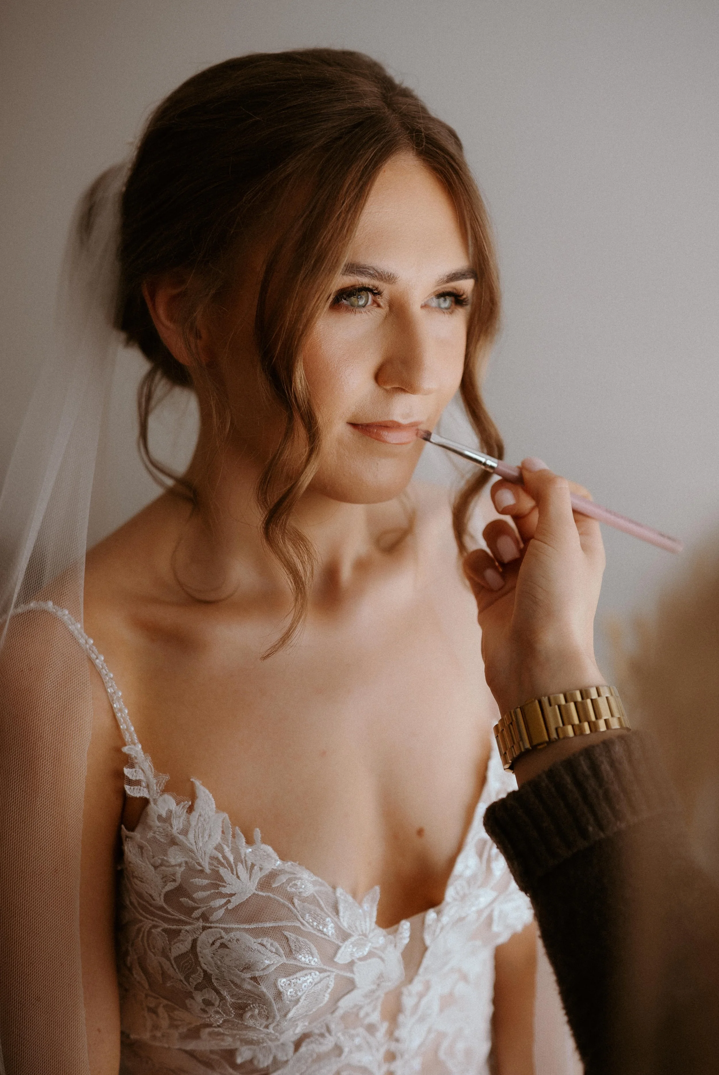 Close-up of a woman with light skin and brown hair styled in loose waves, wearing a white lace dress, receiving makeup application from a hand wearing a gold watch and a dark sweater.