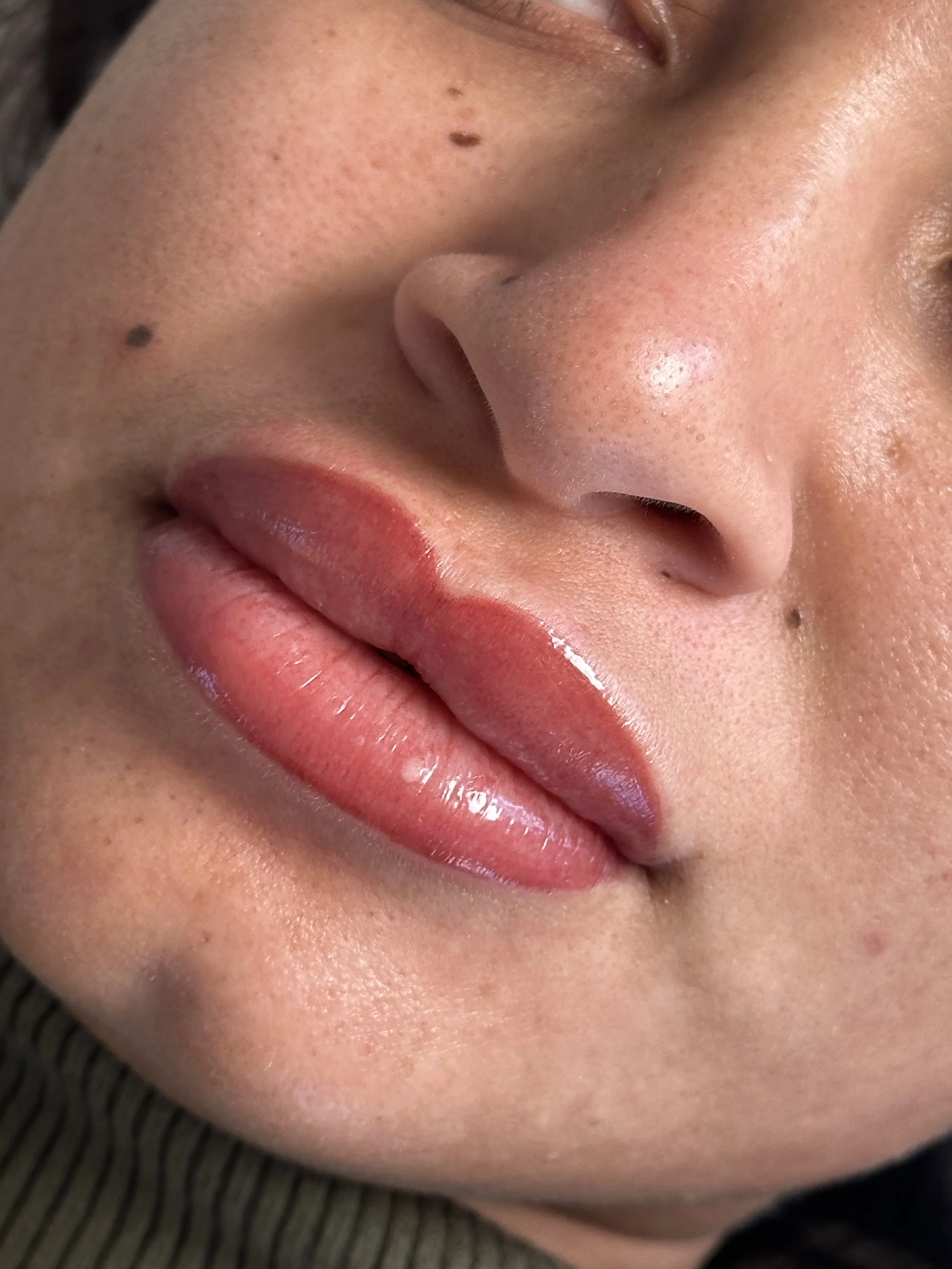 Close-up of a person's face showing lips, nose, and skin with makeup, including lipstick and blush, with moles visible.