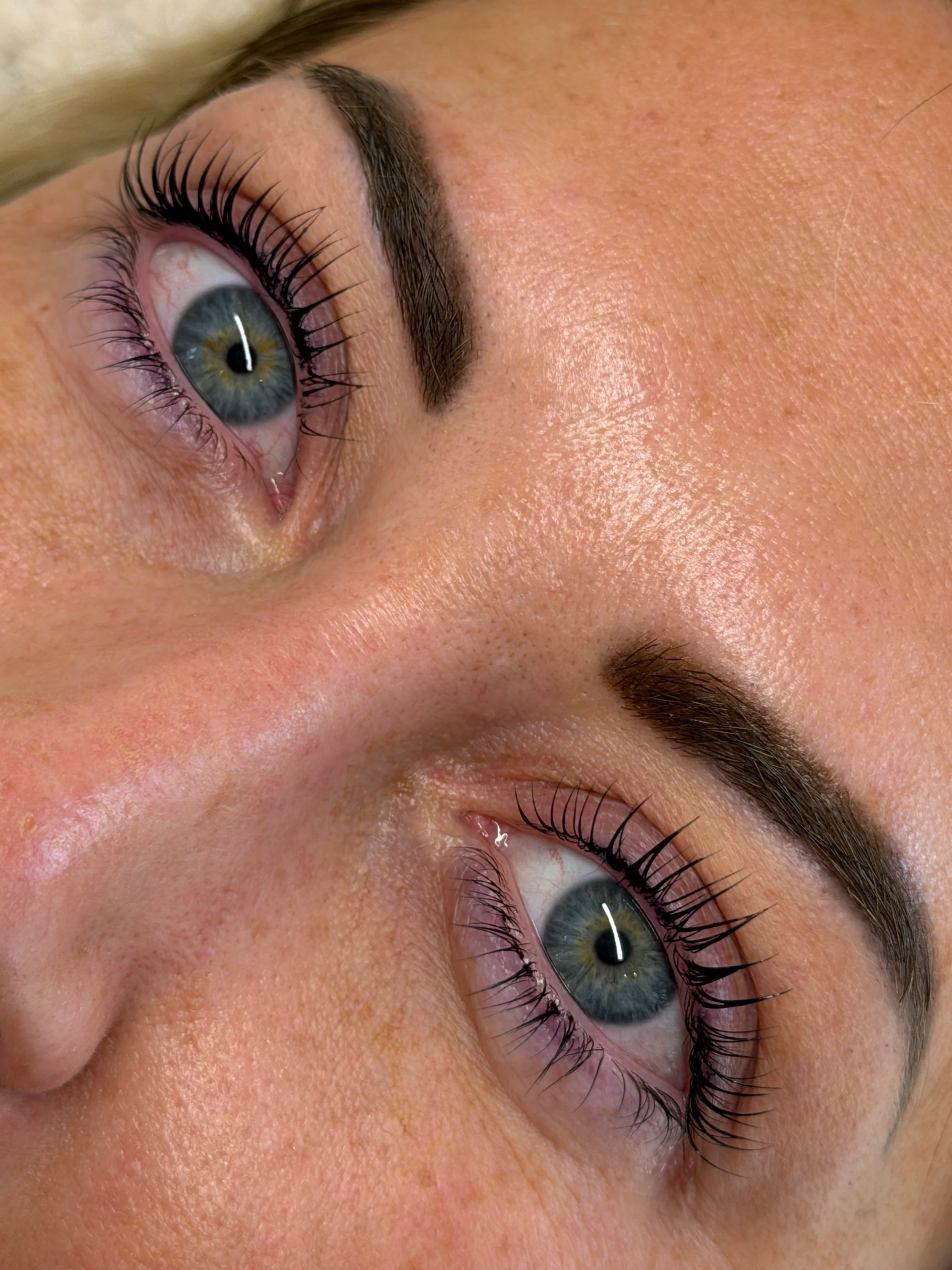 Close-up of a person's face showing blue eyes, long eyelashes, and well-groomed eyebrows, with smooth skin and natural makeup.