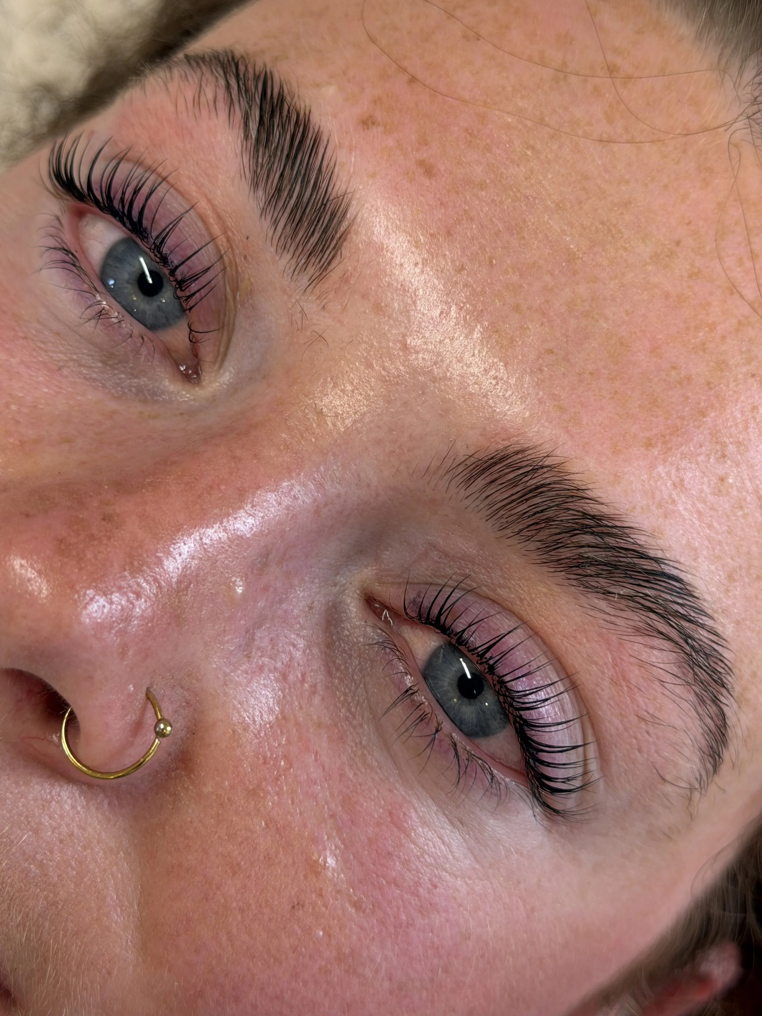 Close-up of a person's face, showing blue eyes and well-groomed eyebrows, with some facial skin freckles and piercings in the nose.