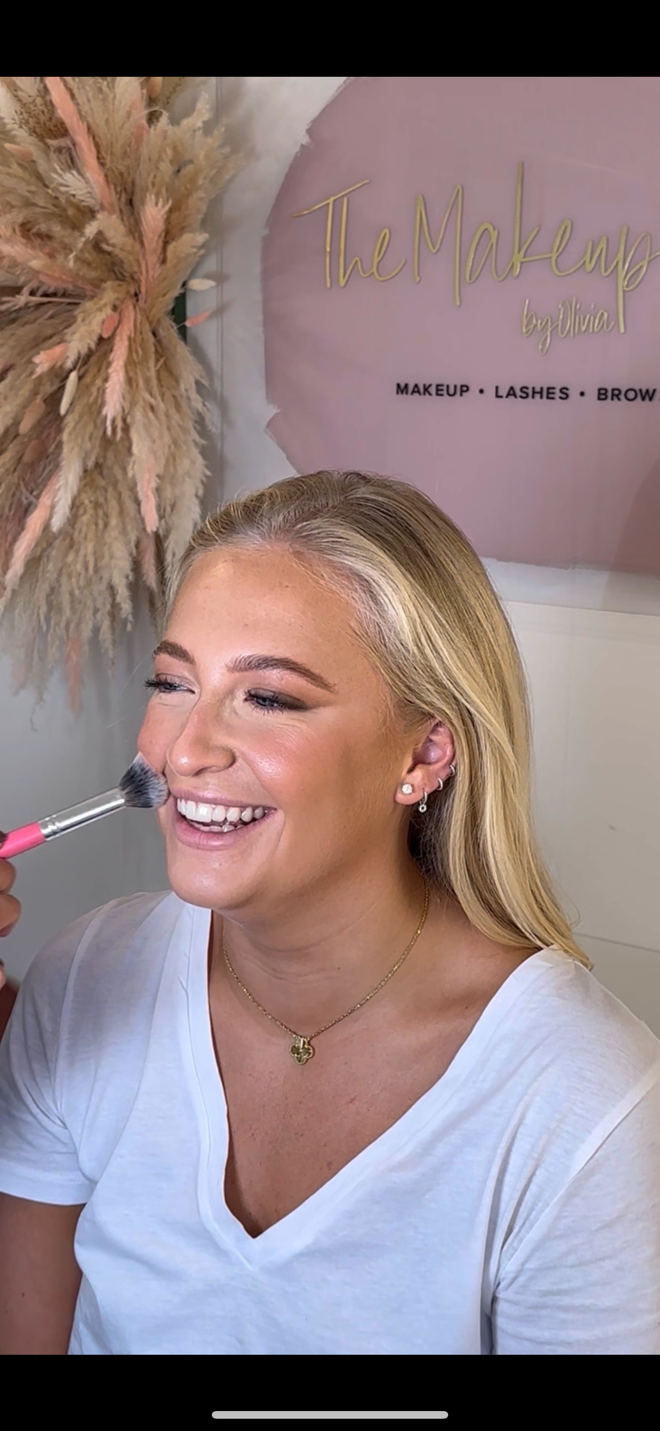 A woman with blonde hair smiling as she gets her makeup done, with a sign in the background that reads 'The Makeup by Olivia' and 'Makeup, Lashes, Brows'.