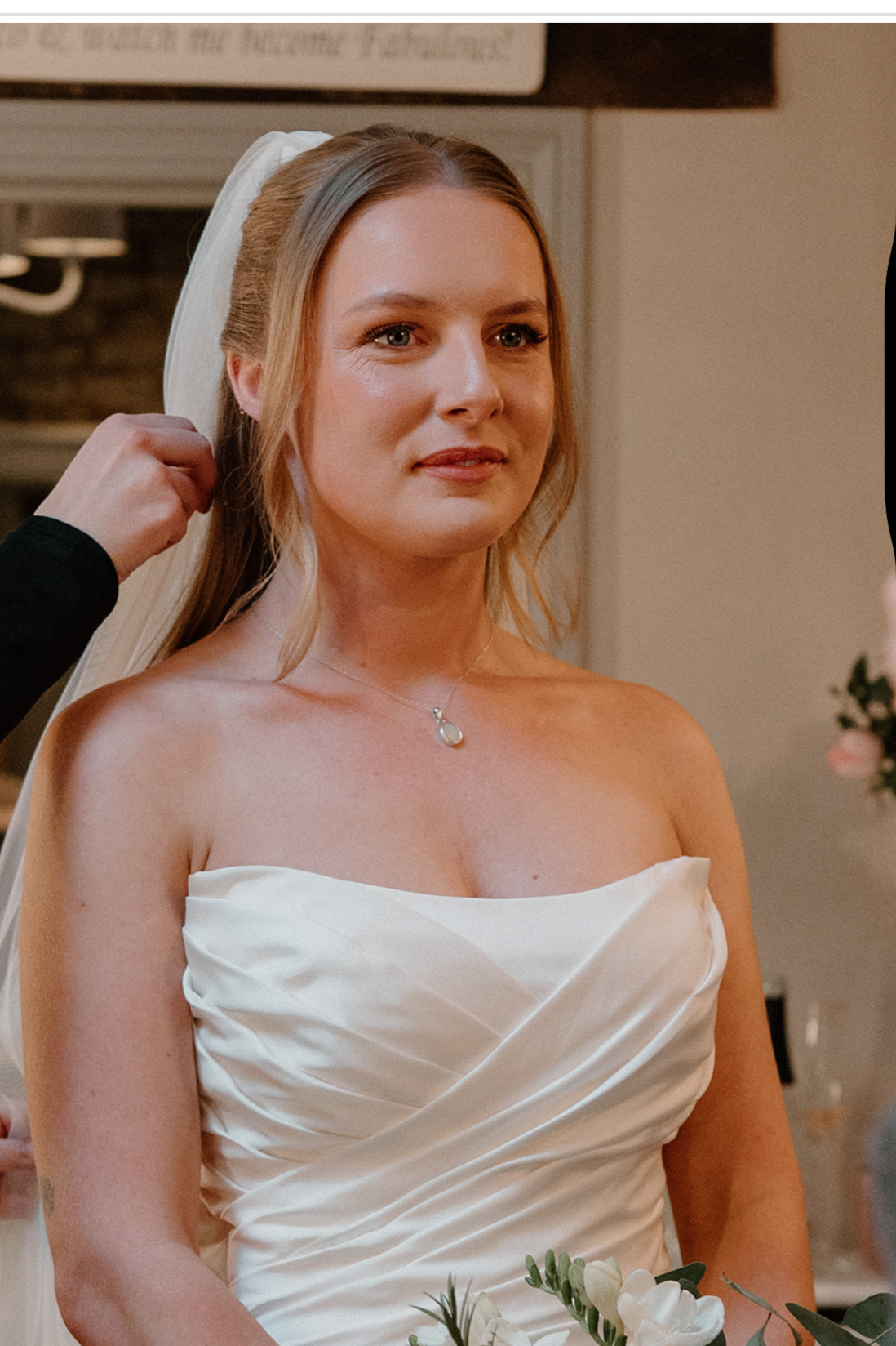 A bride with light skin, wearing a strapless satin wedding dress, a pendant necklace, and a wedding veil, stands indoors with someone adjusting her veil.