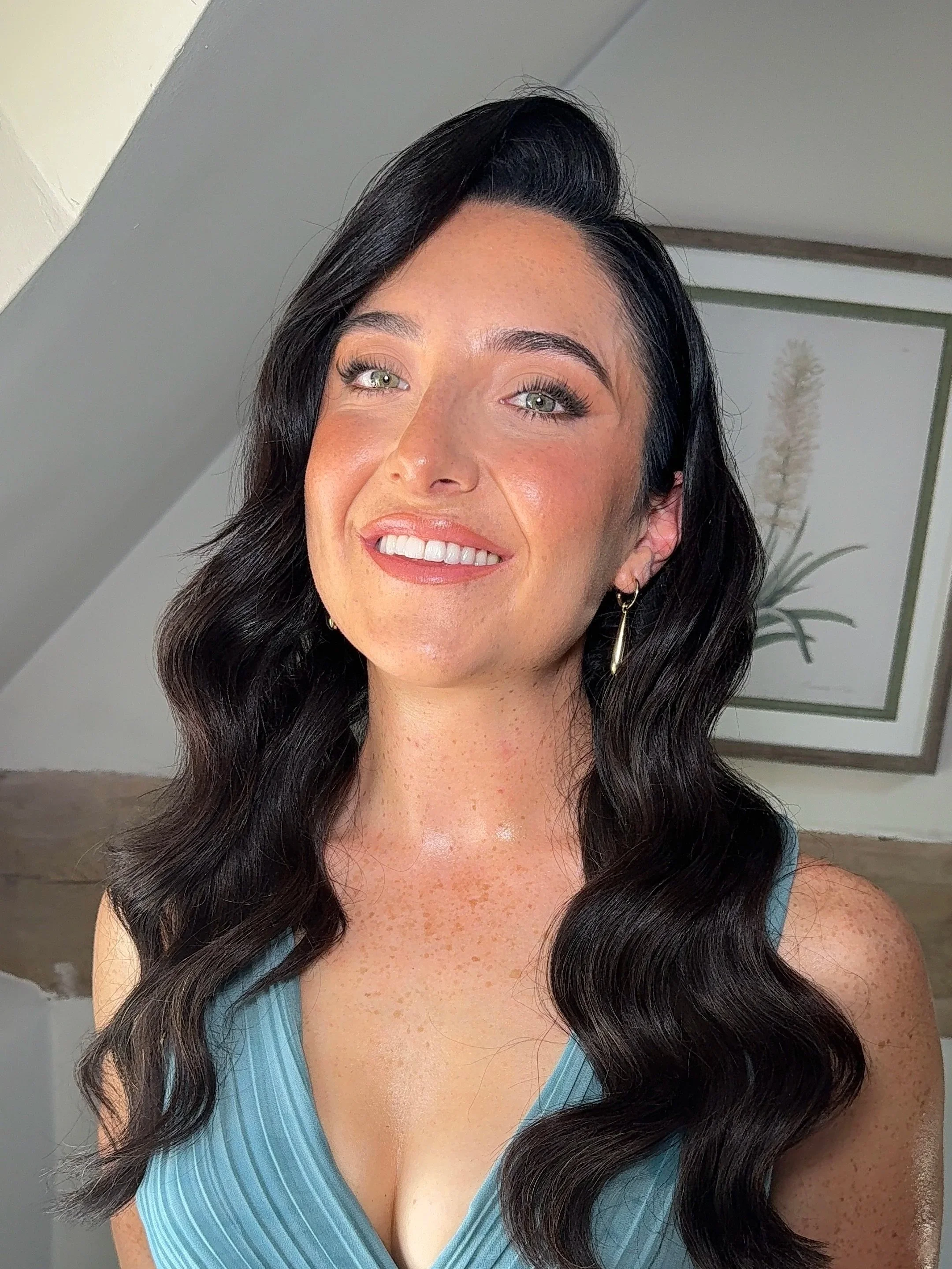 A woman smiling with long dark curly hair and wearing a sleeveless light blue striped top, standing in a room with a framed botanical picture on the wall behind her.