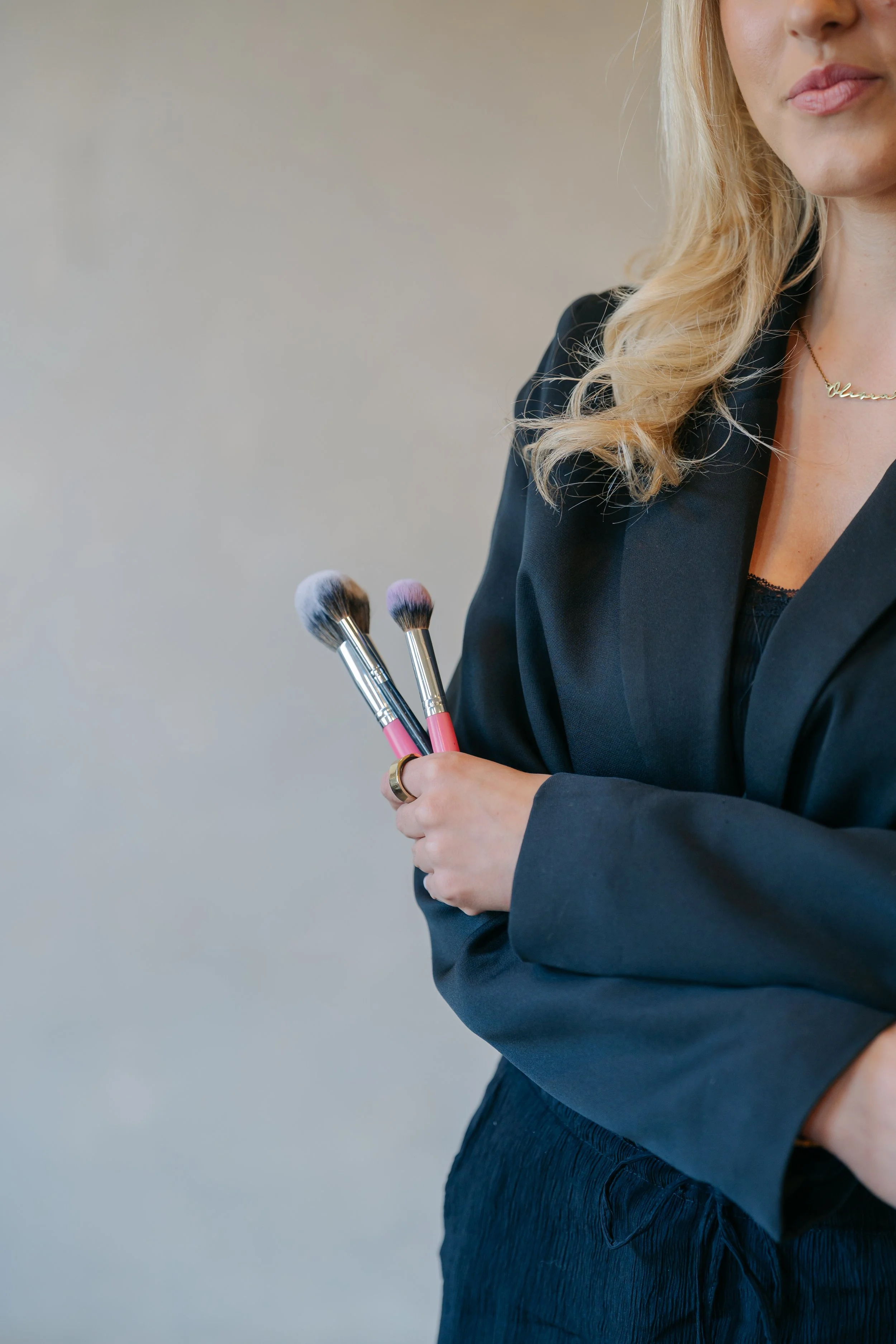 Part of a woman with long blonde hair, wearing a black blazer and gold necklace, holding makeup brushes, with a plain background.