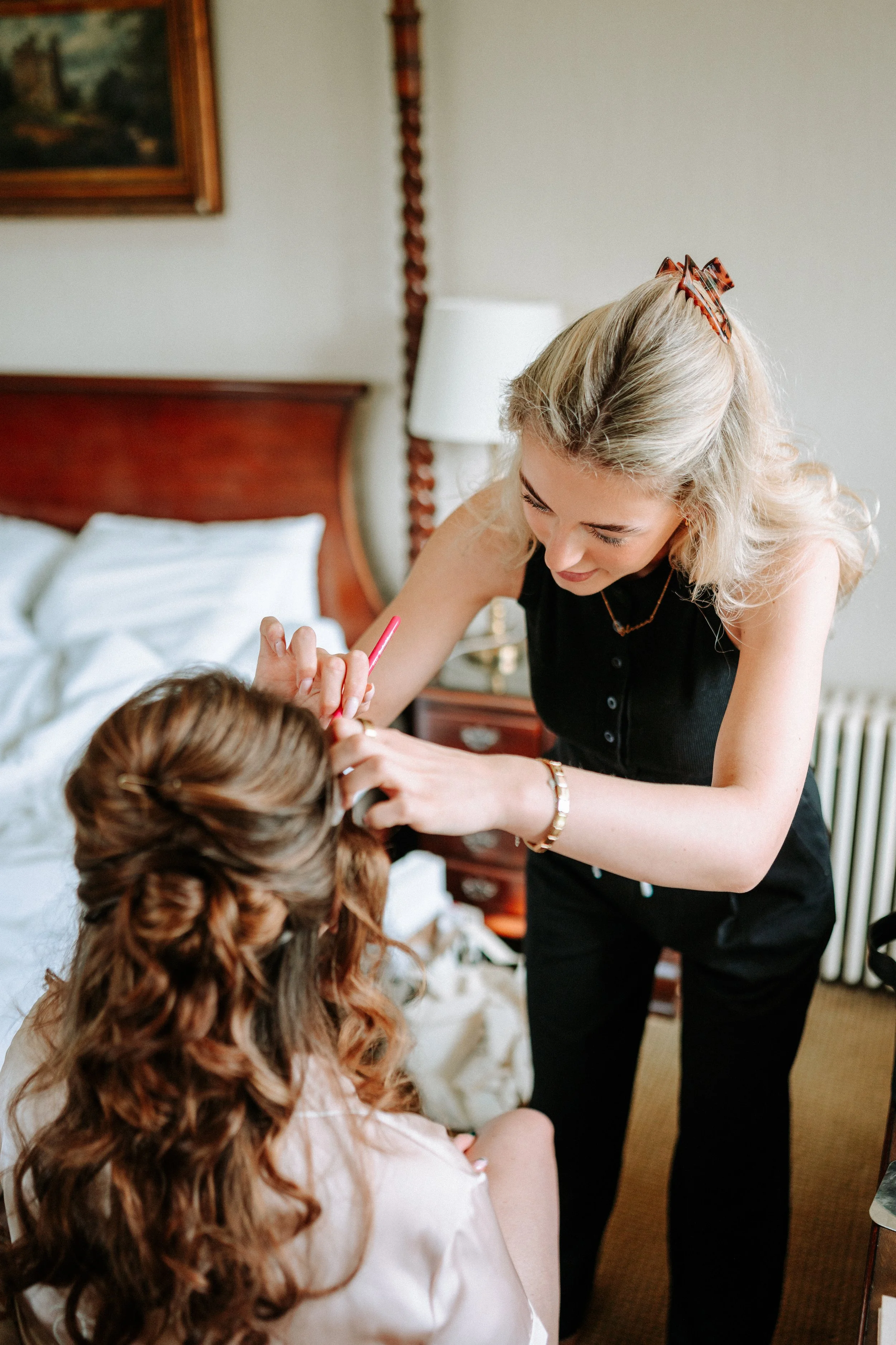 A woman with blonde hair and a red patterned bow hair accessory is applying makeup or lipstick to a young girl with curled brown hair in a bedroom with a bed, pillows, a wooden headboard, an oil painting, a woman in white, and a lamp.