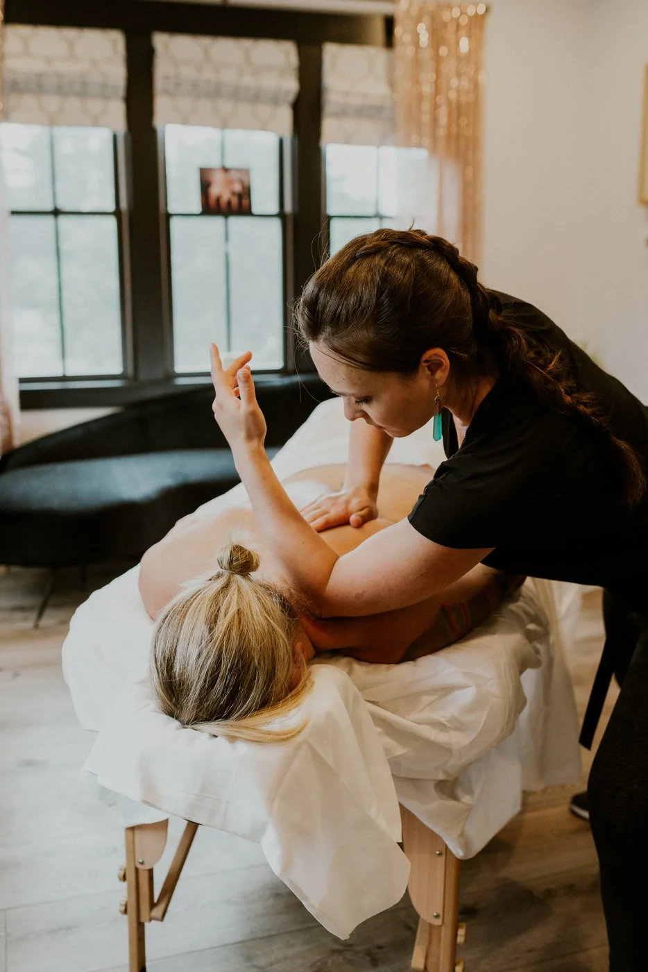 A therapeutic massage therapist uses her elbow to dig deep into a clients shoulder knots. The room has soft lighting and her client is face down, very relaxed.