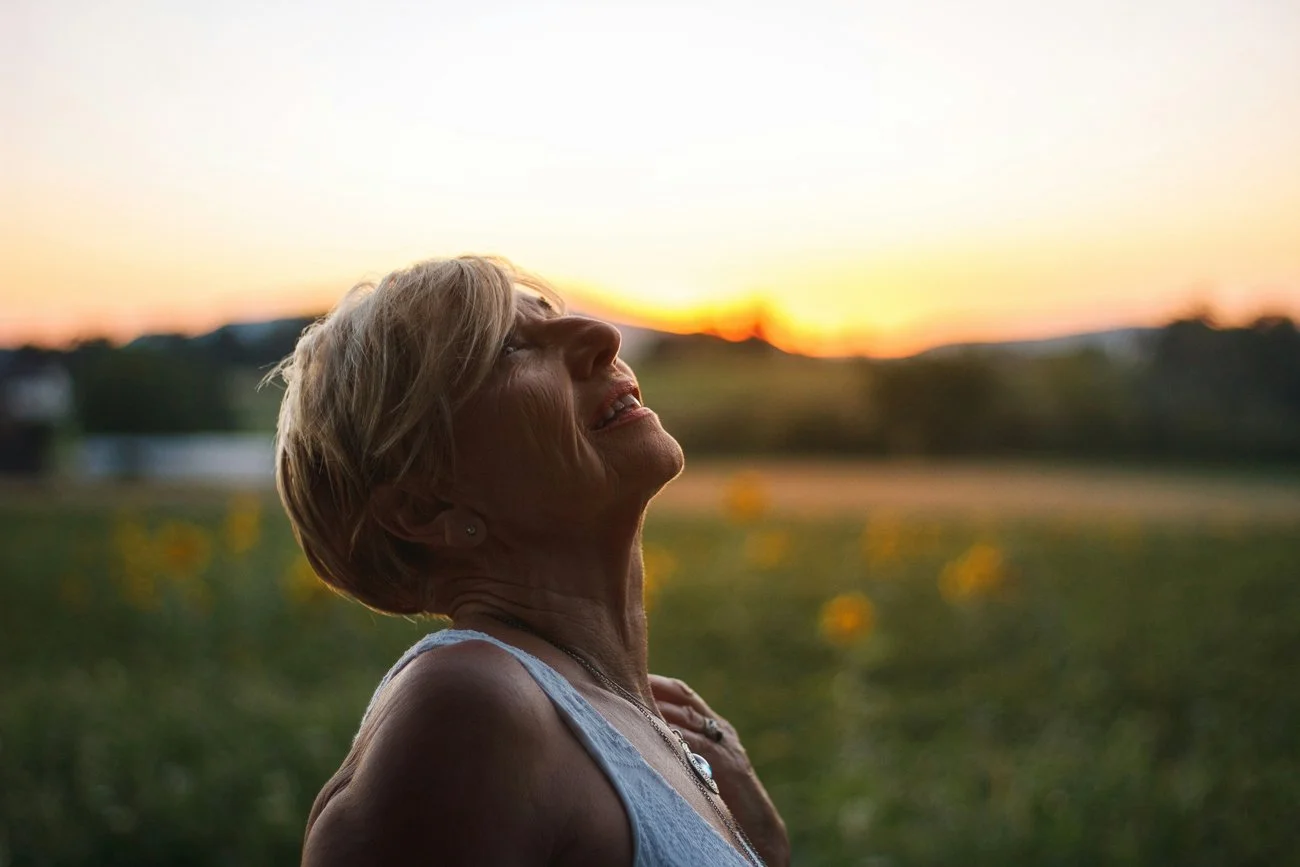 A woman in her fifties looks up at the sky at sunset. She is a client of a health and wellness coaching program that has given her energy and weight loss.