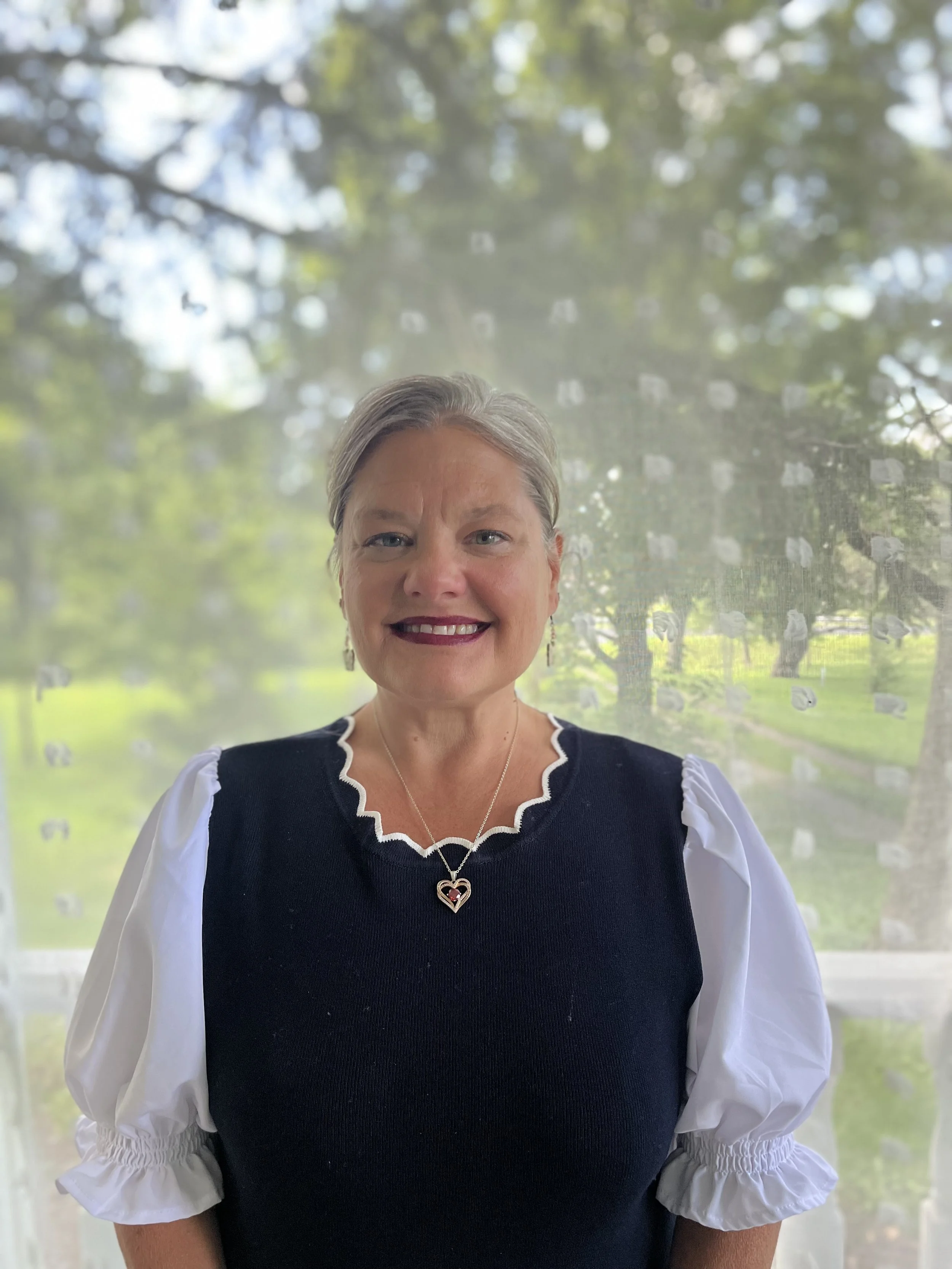 A woman with gray hair wearing a black top with white puffed sleeves and a white shirt underneath, standing outdoors with a blurred background of trees and greenery.
