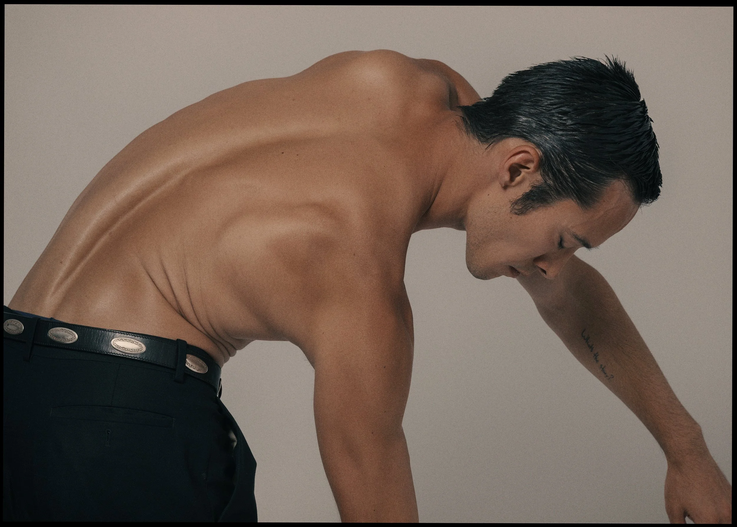 Portrait of a shirtless male model with wet hair leaning forward in a sculptural pose against a neutral studio background.