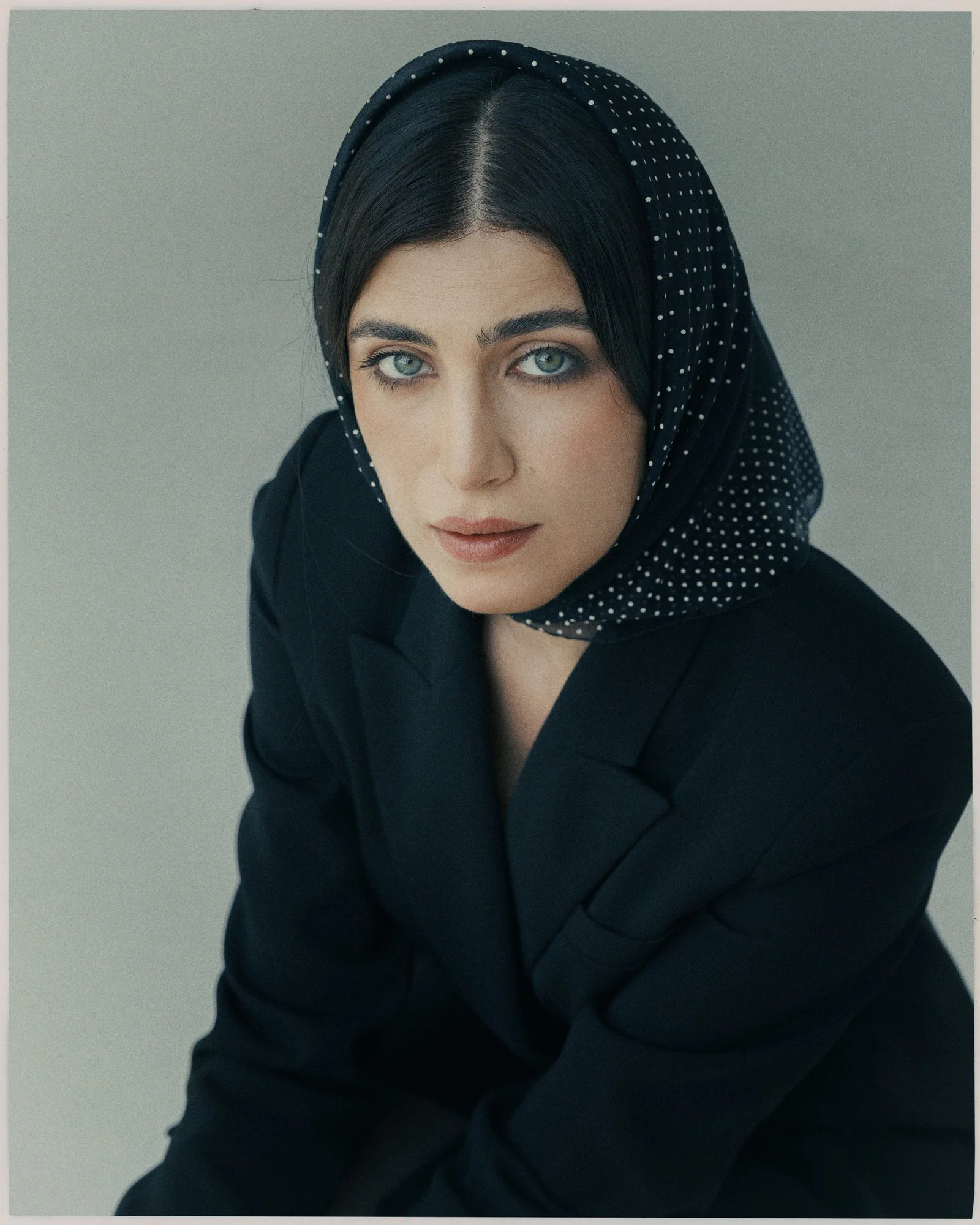 A woman with dark hair, hazel eyes, and light skin posing for a portrait against a plain gray background, wearing a black headscarf with white polka dots and a black blazer.