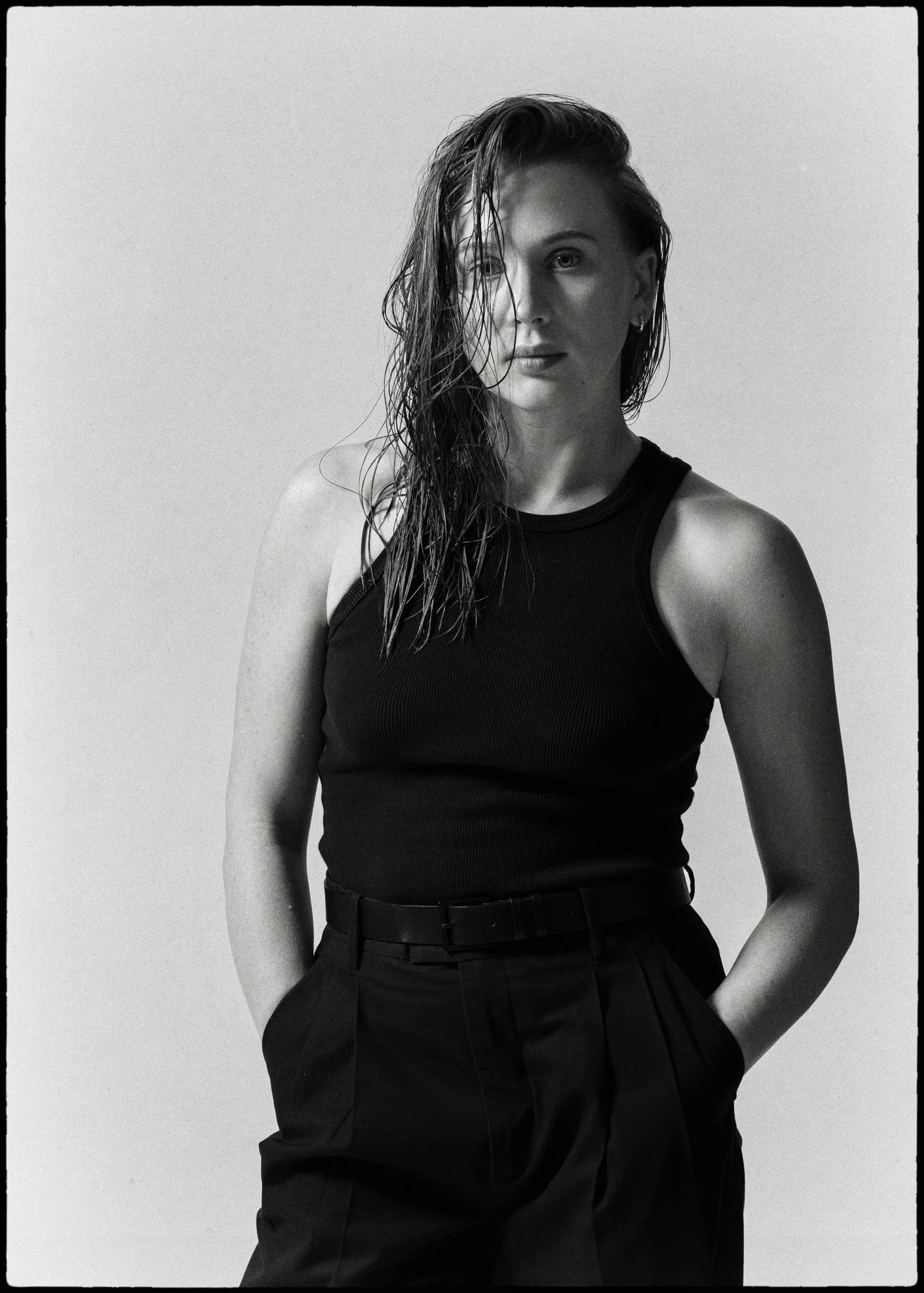 A woman with wet, wavy hair wearing a black tank top and black high-waisted pants standing against a plain background.
