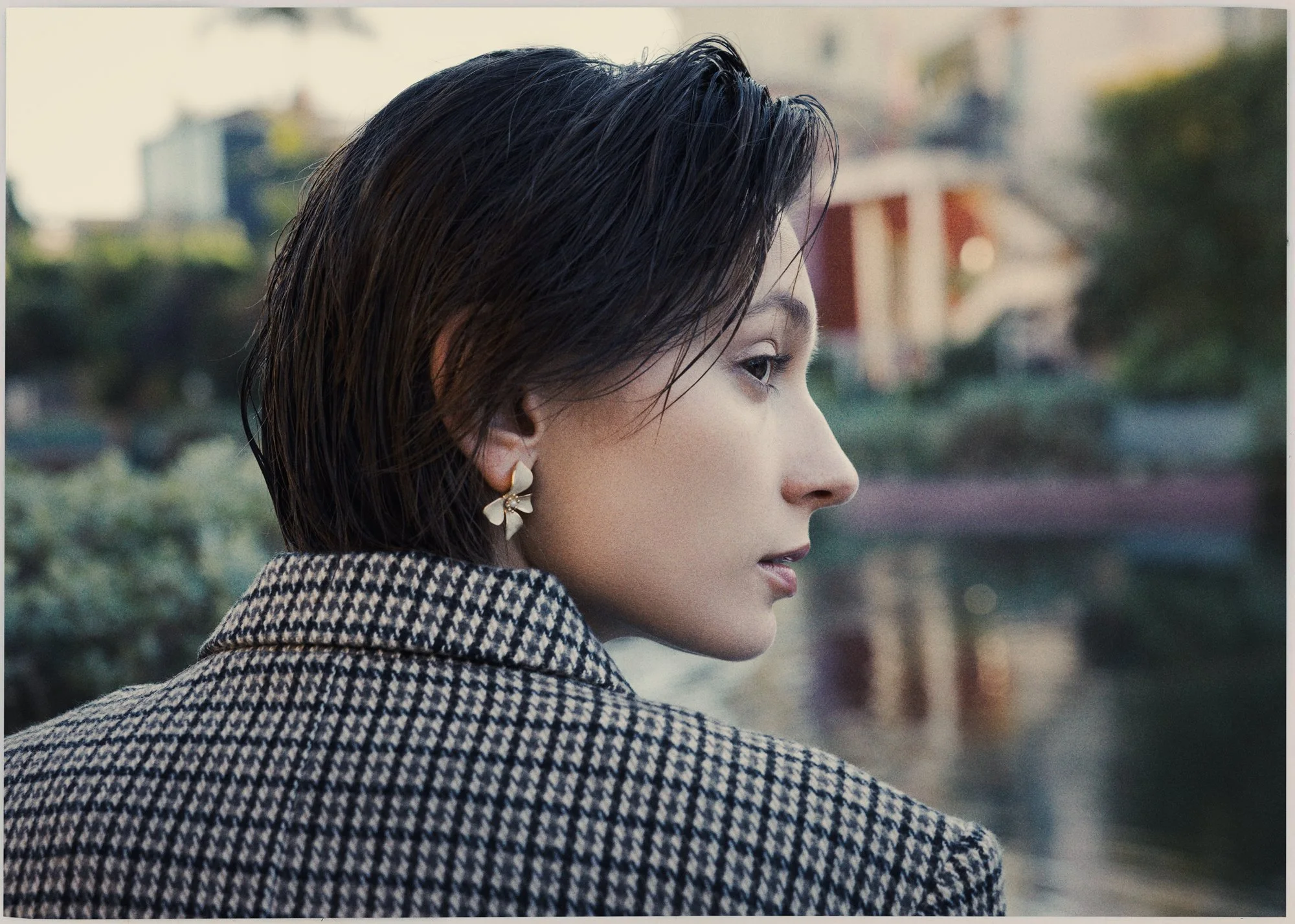 Close-up profile shot of a woman with dark, wet hair, wearing floral earrings and a houndstooth coat, outdoors with blurred greenery and buildings in the background.