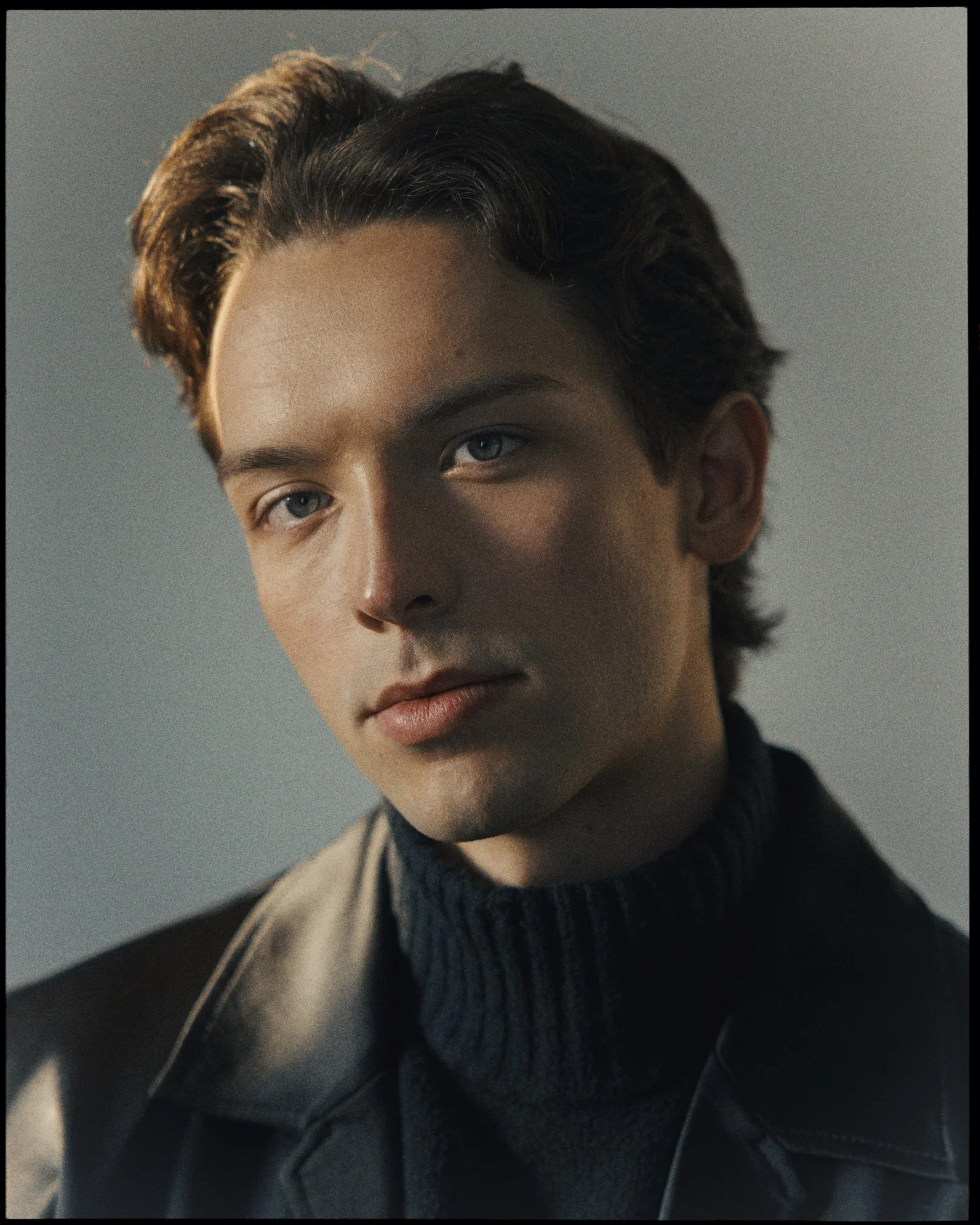 Close-up of a young man with dark, wavy hair and blue eyes, wearing a black turtleneck sweater and a beige coat, in a neutral indoor setting.