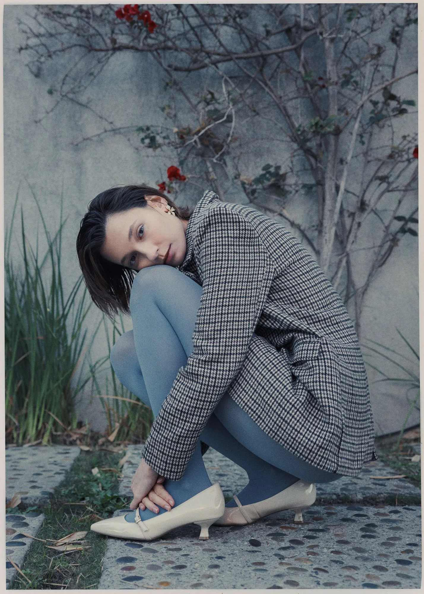 A woman in a checkered blazer and gray leggings tying her beige and blue high heel shoe outdoors on a stone pathway, with tall grass and a tree with red flowers in the background.