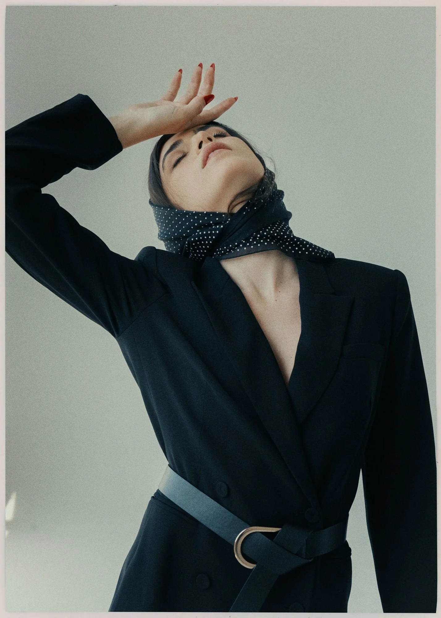 A woman with dark hair, wearing a black blazer and a black scarf with white polka dots, posing with her eyes closed, hand on her forehead, against a light background.