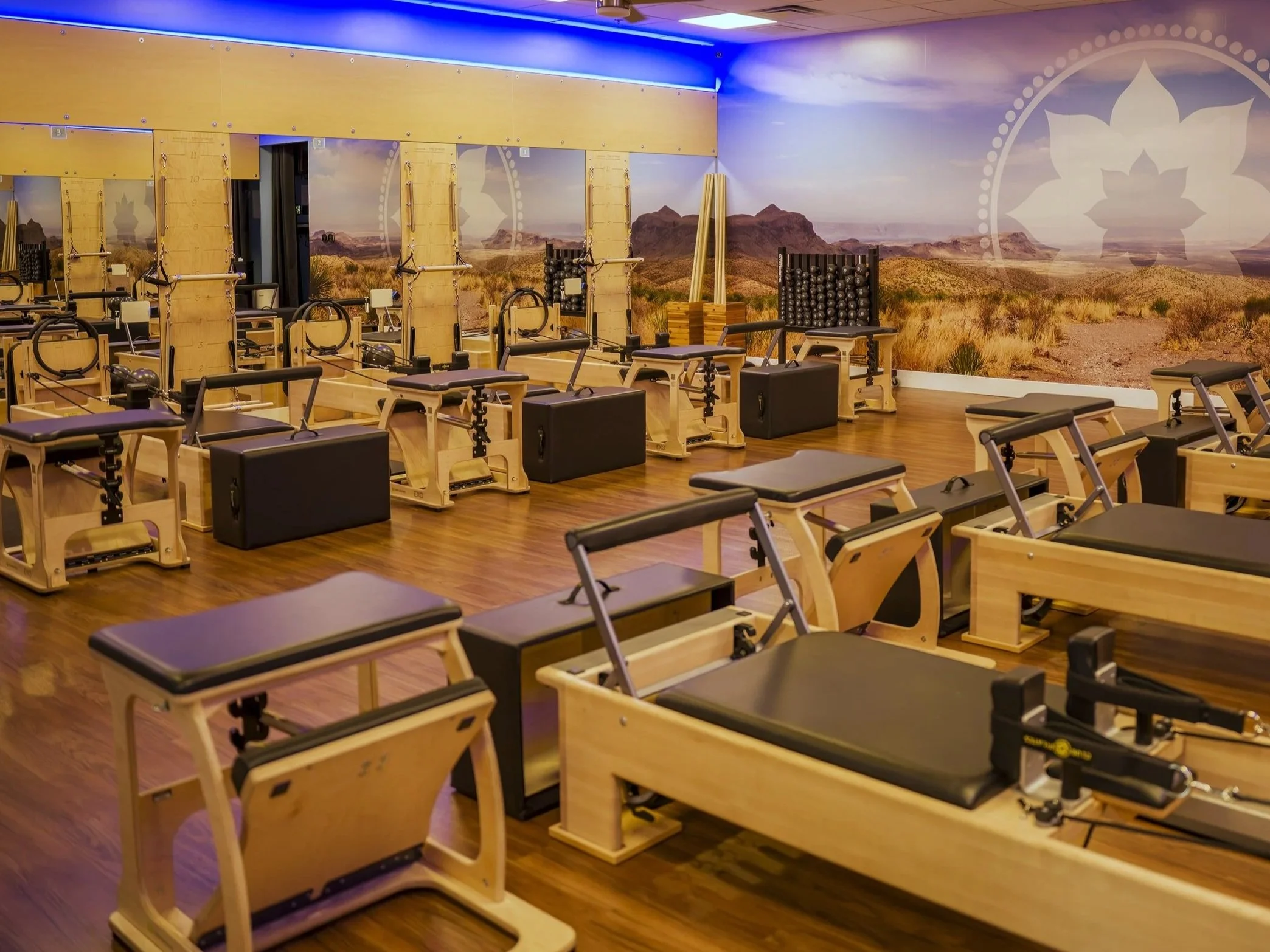 A Pilates or fitness studio with wooden equipment, including reformer machines, in front of a desert landscape mural.