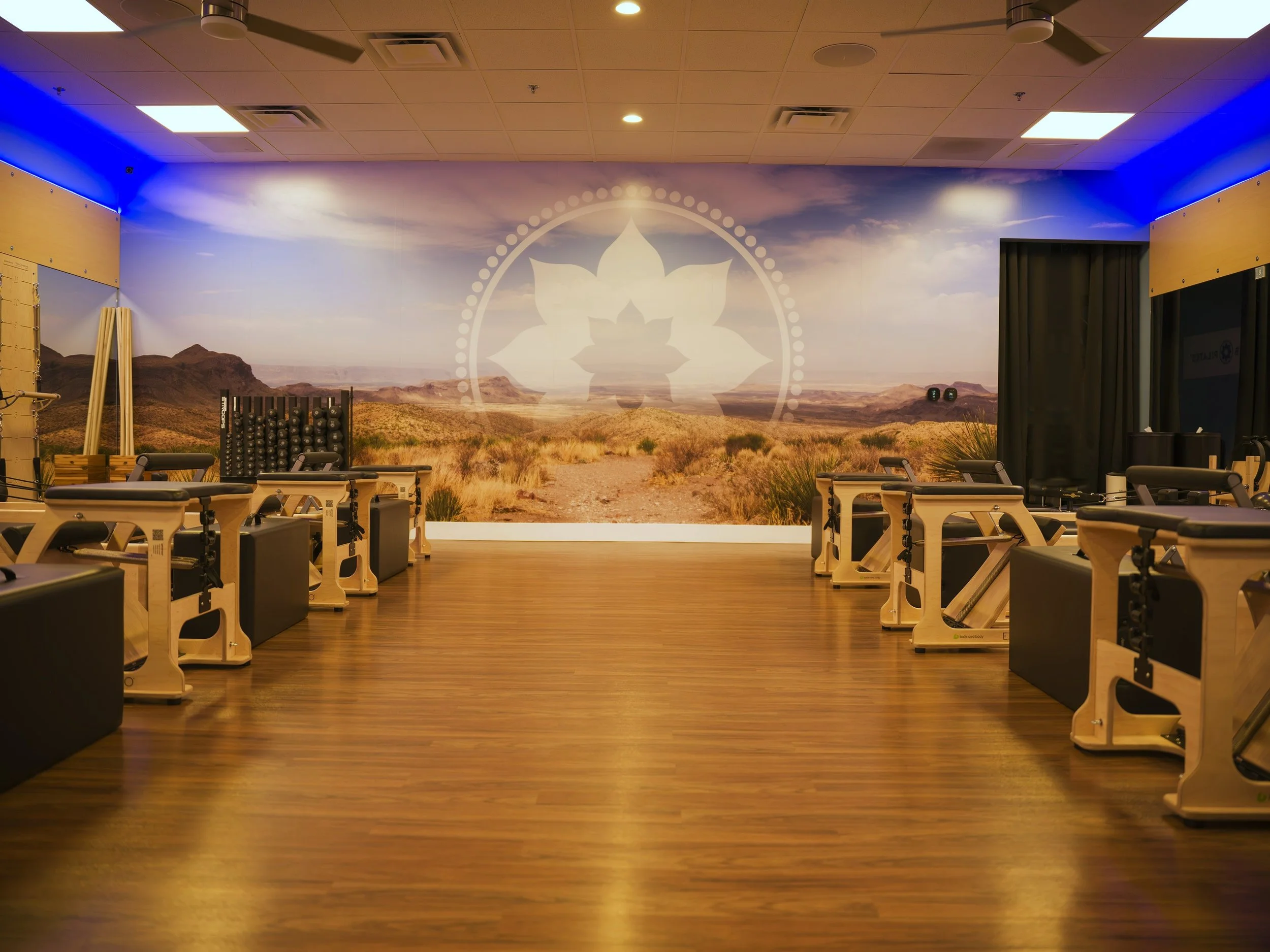 Indoor fitness studio with wooden flooring, workout equipment, and a large mural of a desert landscape with mountains and a blue sky on the wall, featuring a white lotus flower emblem.