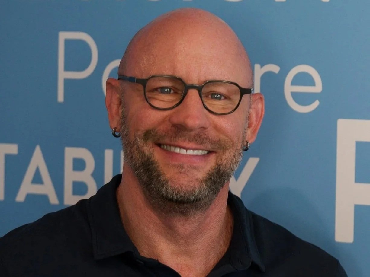 A smiling man with a bald head, glasses, earrings, and a beard, standing in front of a blue background with partially visible white text.