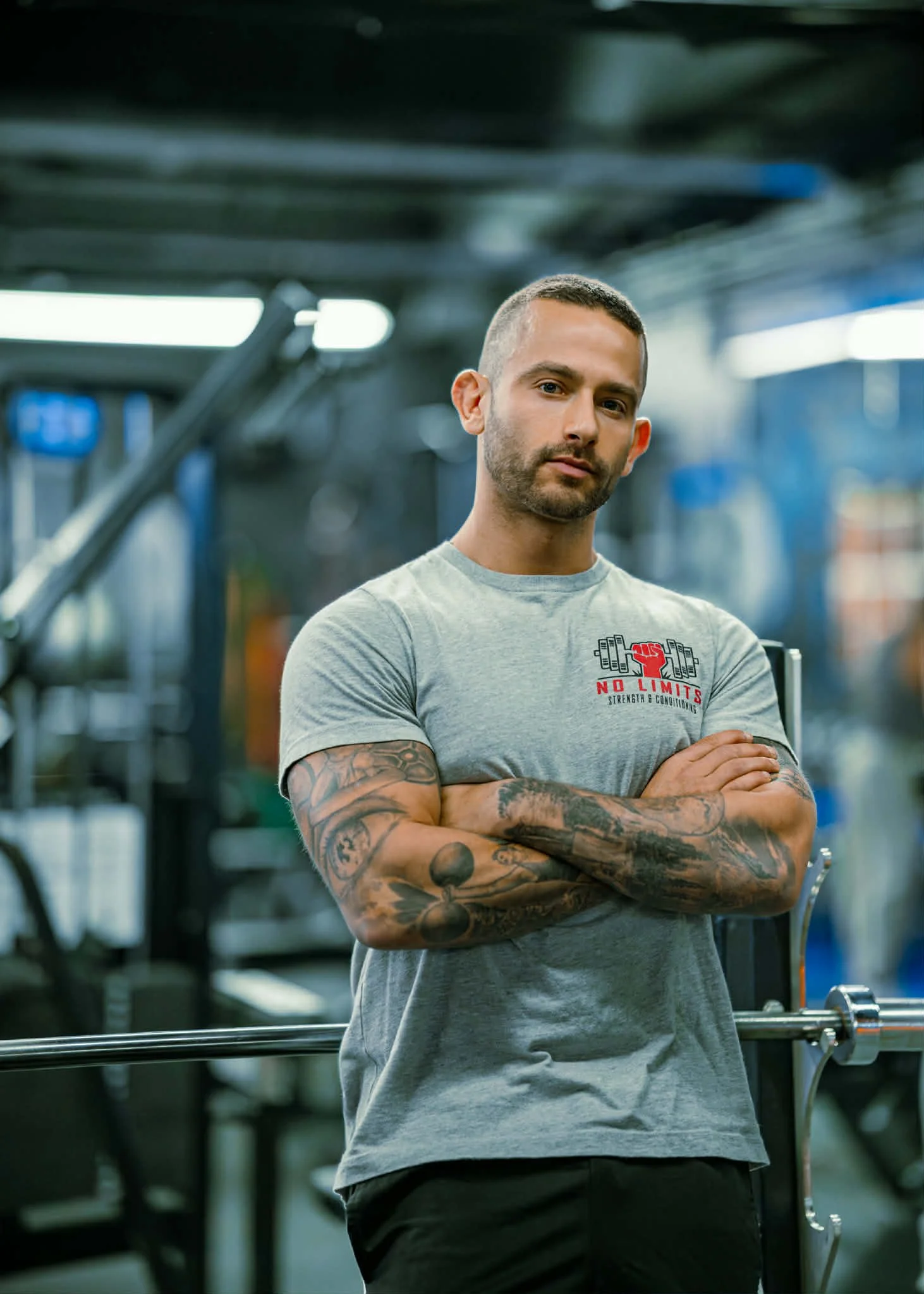 A muscular man with faded hair and tattoos on his arms, wearing a gray T-shirt with a weightlifting logo, standing with his arms crossed in a gym.