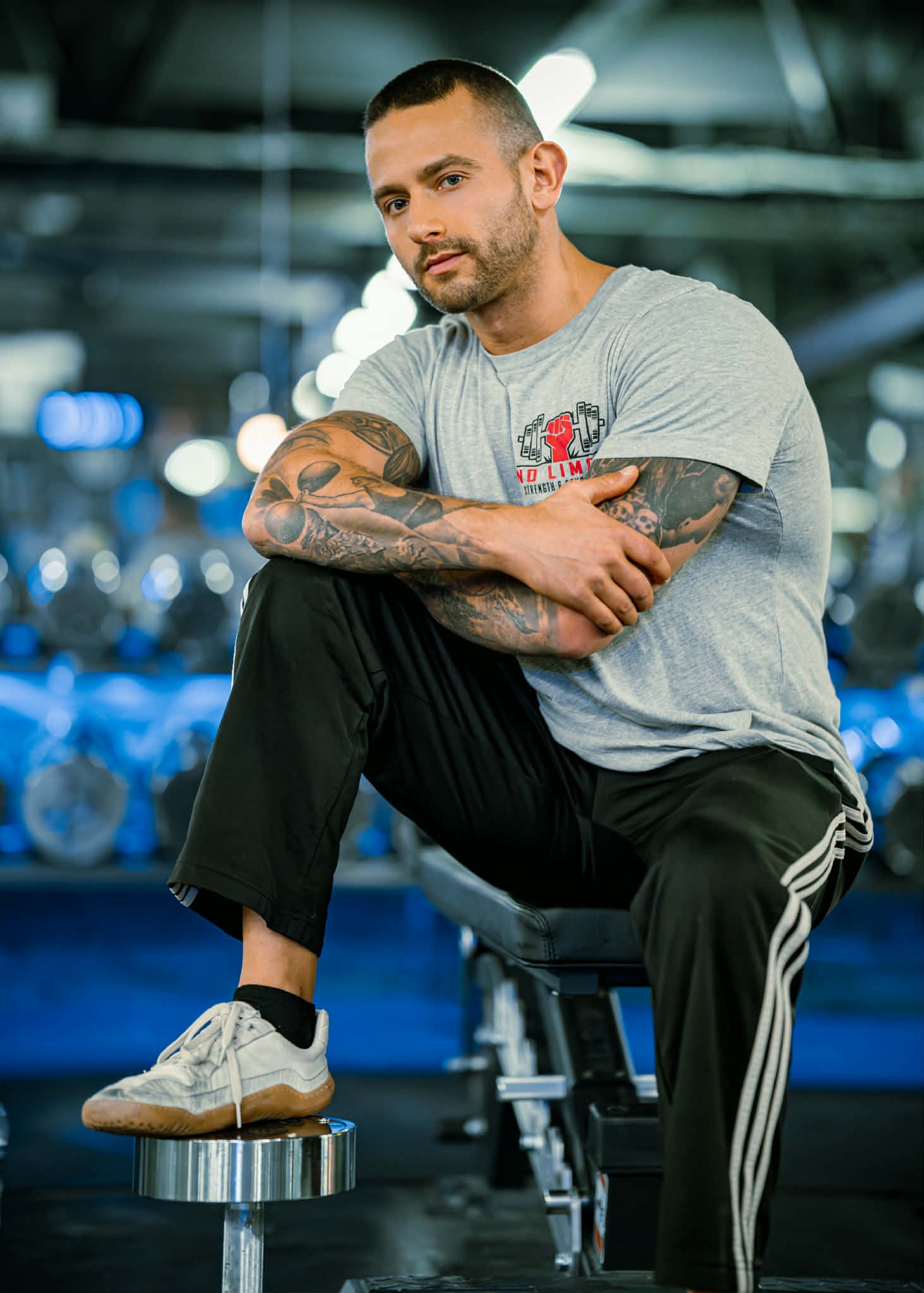 A young man with tattoos, wearing a light gray t-shirt and black track pants with white stripes, sitting on a workout bench in a gym, Brady's left elbow resting on his right knee, looking at the camera.