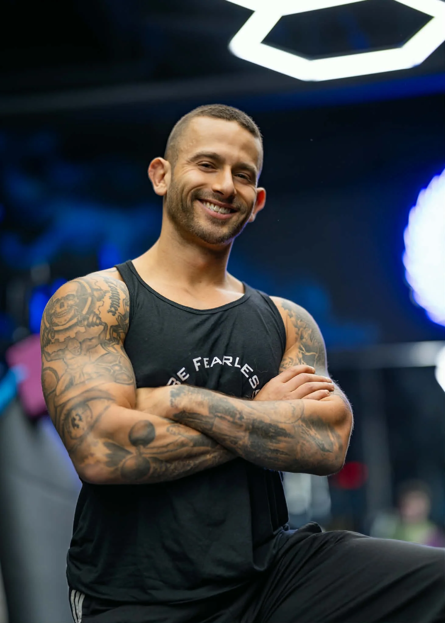 Smiling man with short hair and tattoos on his arms, wearing a black tank top, sitting with arms crossed in a gym.