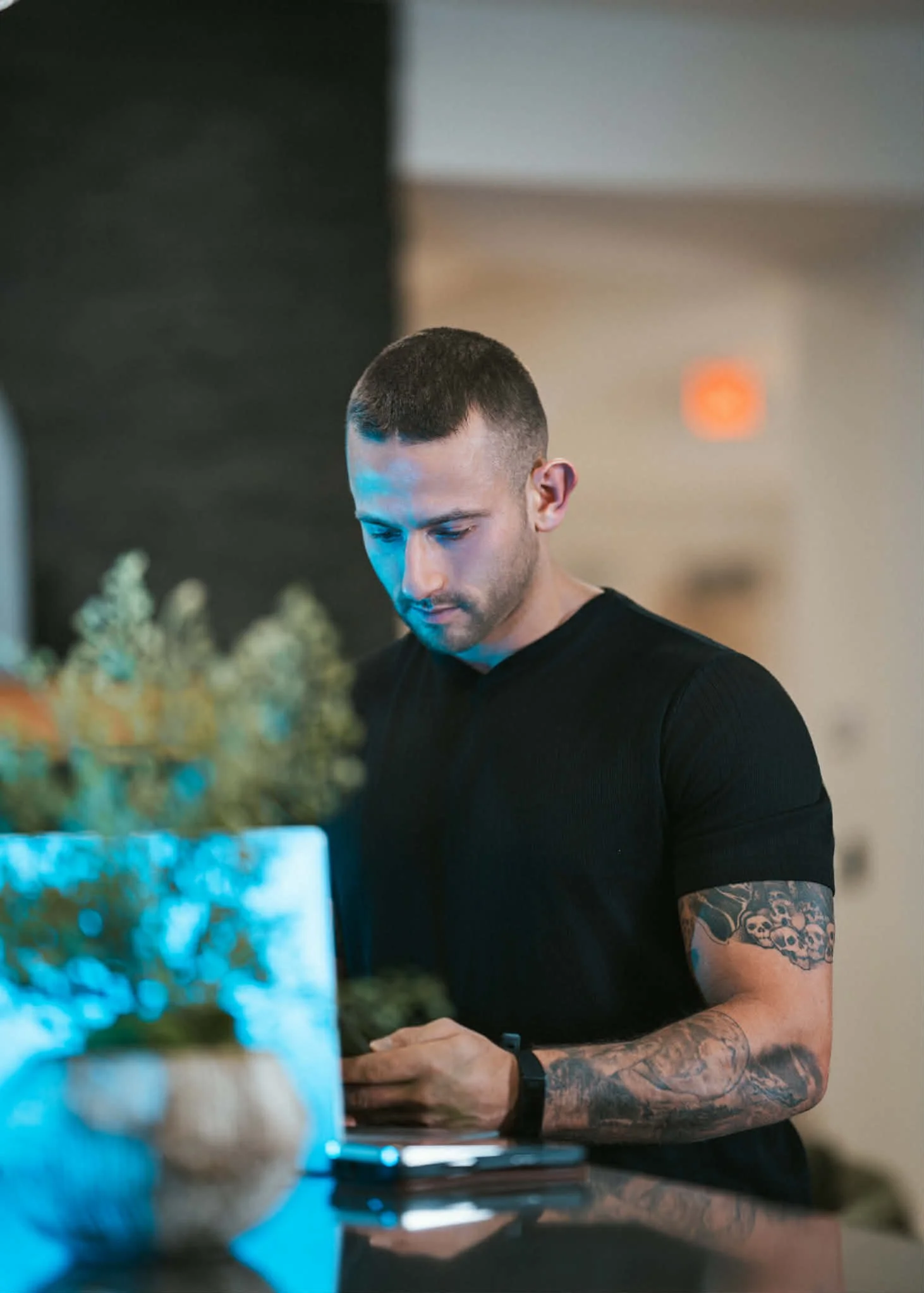 A young man with tattoos on his arms, short hair, and wearing a black t-shirt, is standing at a counter, looking down at a phone in his hand. The background is blurred with a neutral wall and a small orange light visible.