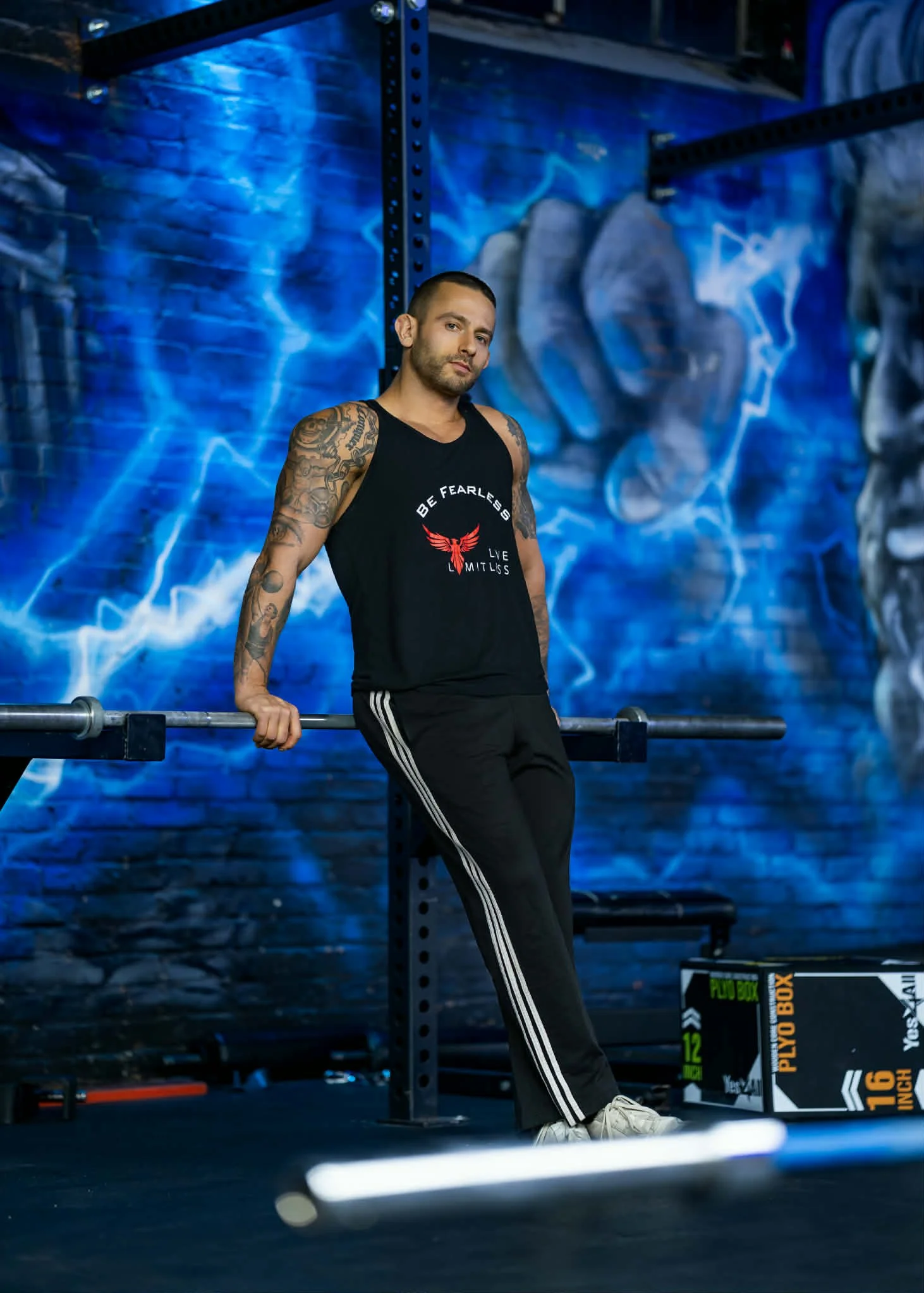 A man with tattoos wearing a black sleeveless shirt and black pants with white stripes, standing in a gym with blue lightning-themed wall art, leaning on a barbell.