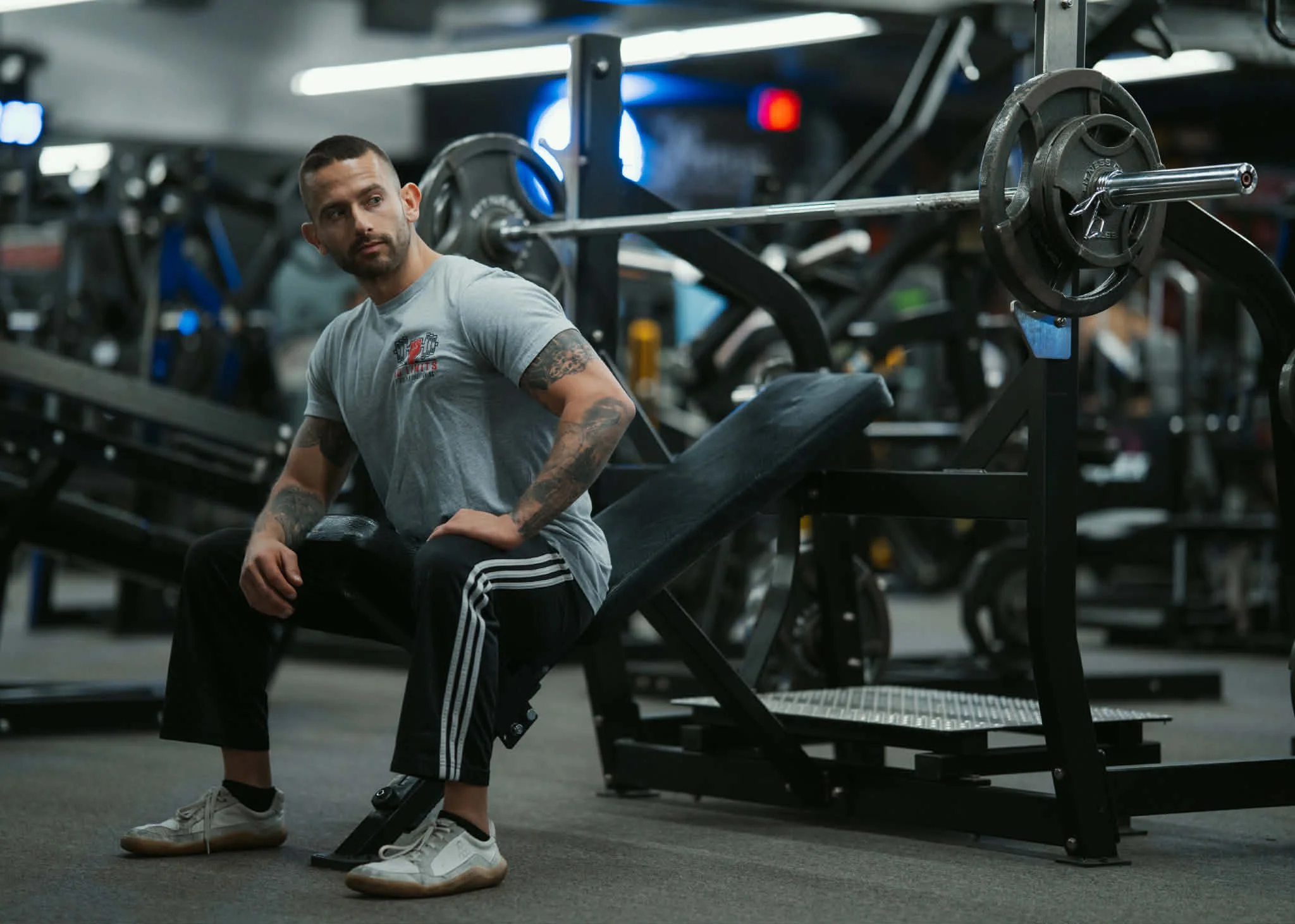 A man with tattoos on his arms wearing a gray T-shirt and black Adidas pants, sitting on a bench inside a gym with weightlifting equipment in the background.