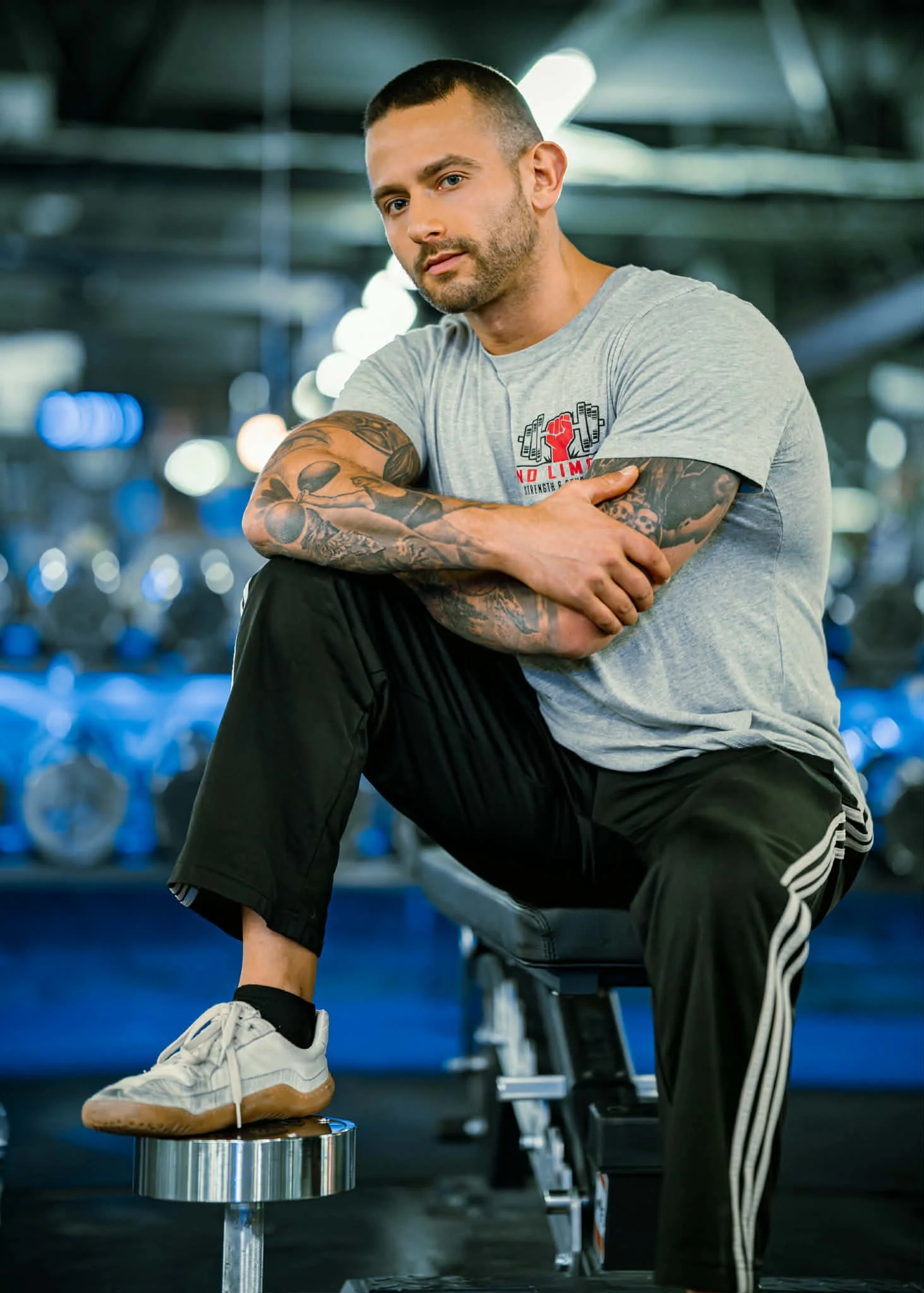 A man with short dark hair and tattoos on his arms, wearing a light gray t-shirt and black athletic pants, sitting on a workout bench in a gym with bright lights and rows of dumbbells in the background.